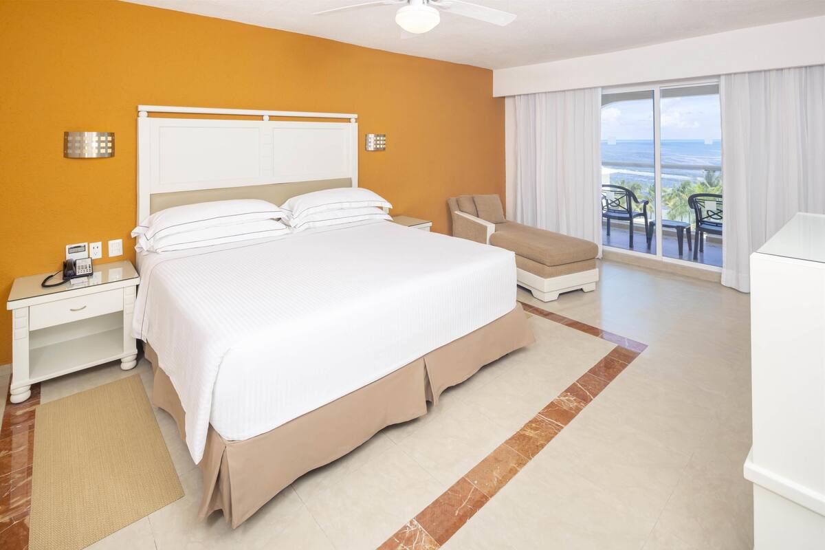 Habitación de hotel con una cama grande, un escritorio con teléfono, una silla y vista al océano desde un balcón.