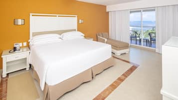 Een hotelkamer met een groot bed, een bureau met telefoon, een stoel en uitzicht op de oceaan vanaf een balkon.