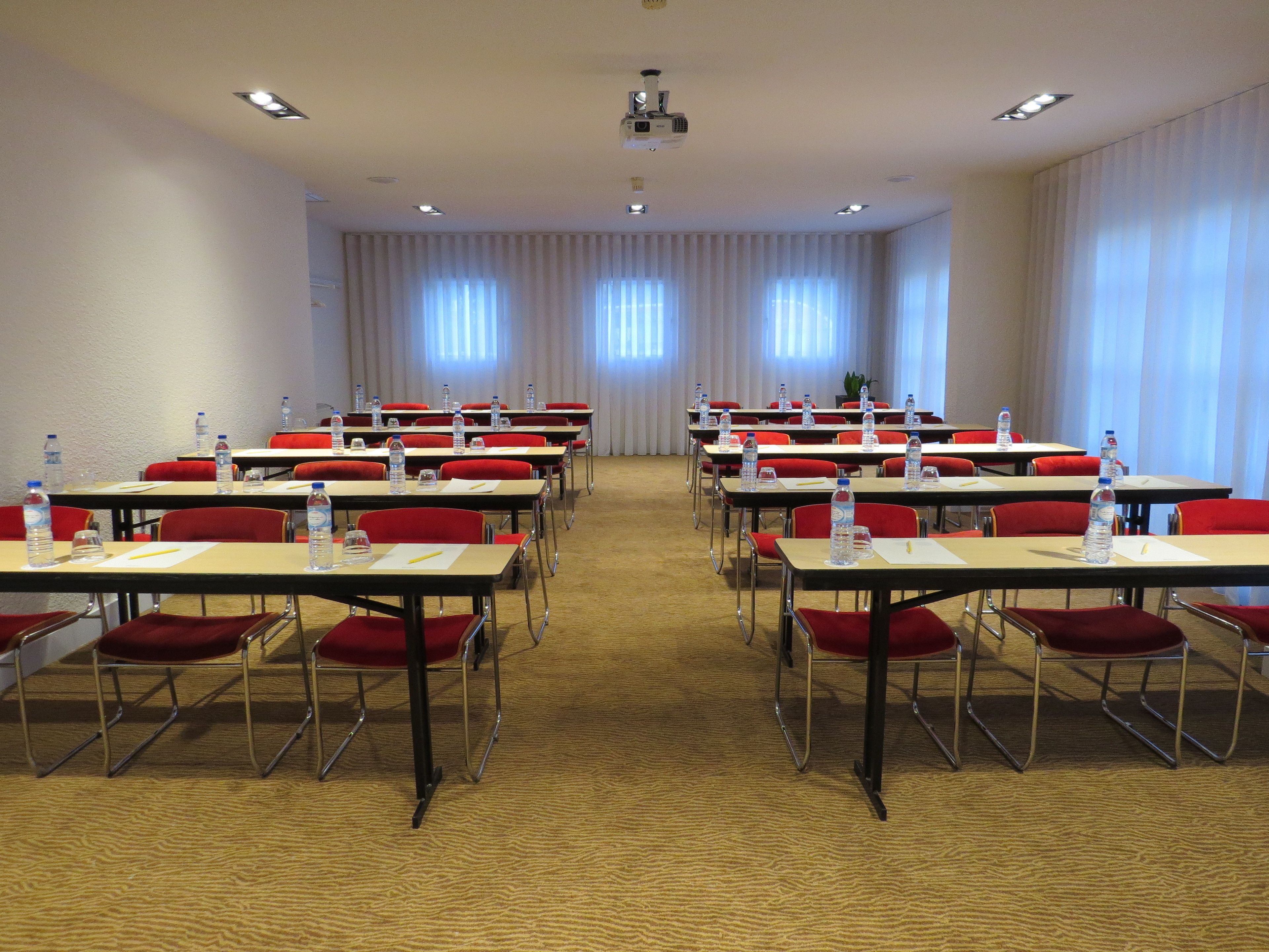 Una sala conferenze con file di tavoli e sedie, dotata di bottiglie d'acqua e documenti, e un proiettore nella parte anteriore.