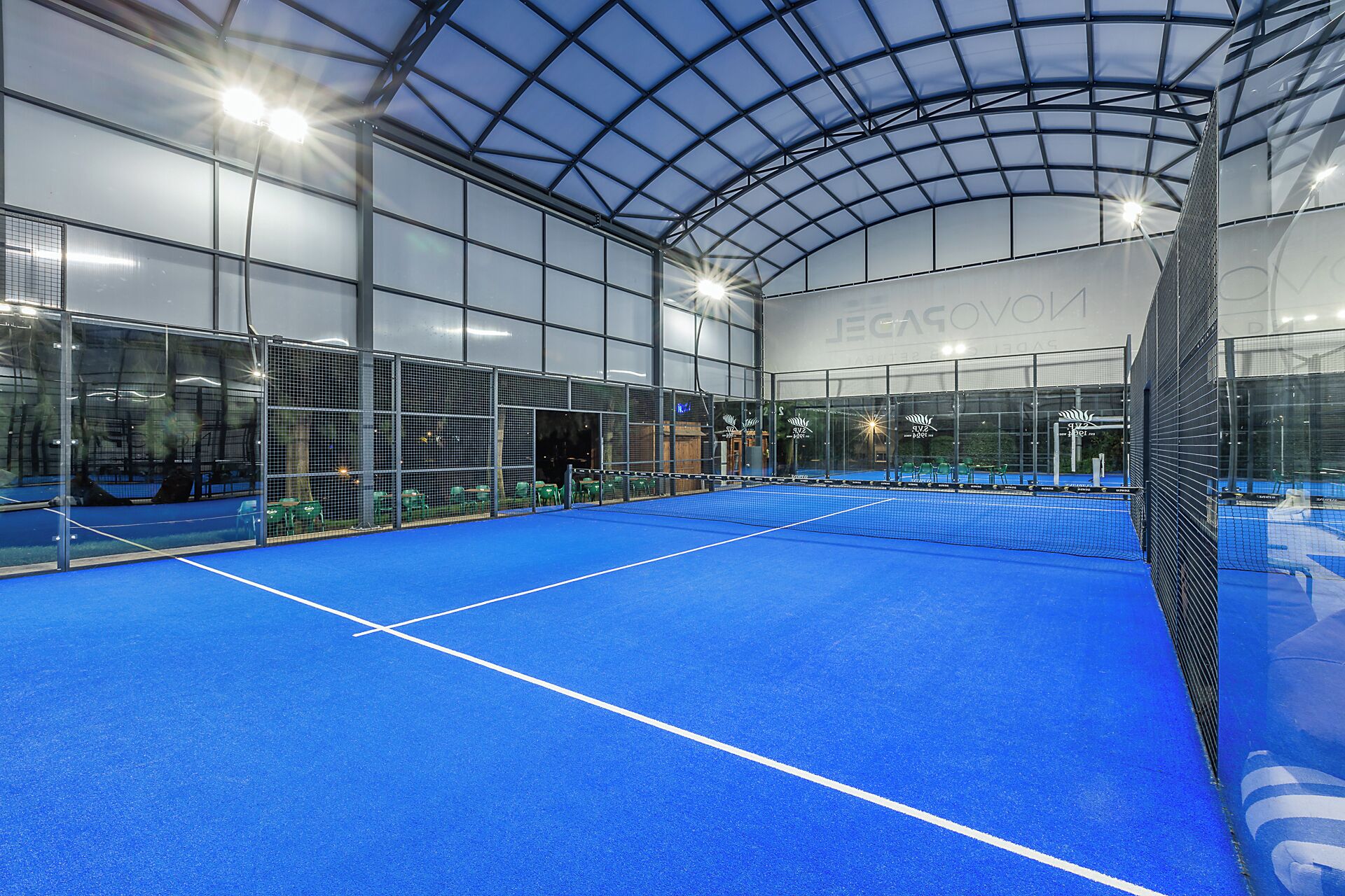 Una cancha de tenis cubierta con superficie azul, rodeada por paredes de vidrio y una estructura de techo metálico.