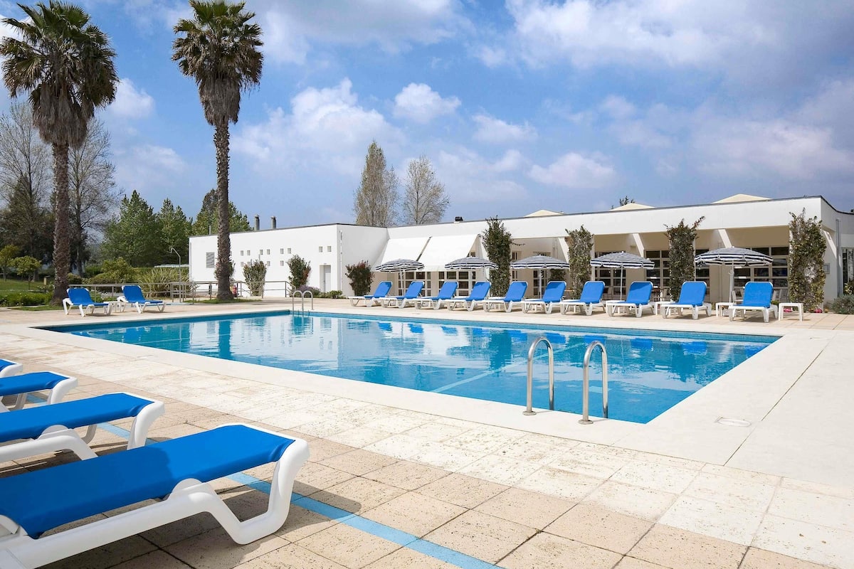 Une piscine bordée de chaises longues bleues et d’ombrelles, entourée de palmiers et d’un bâtiment à la façade blanche.