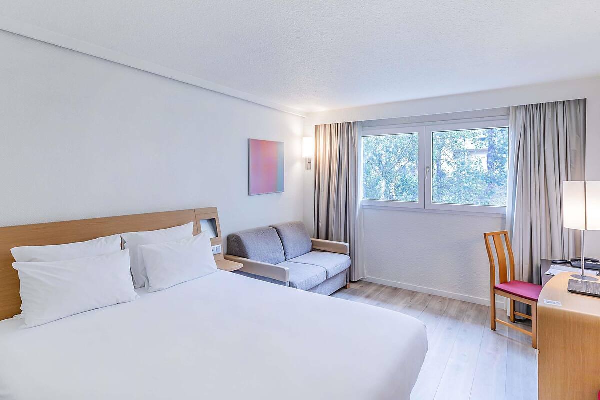 Habitación de hotel con cama, sofá, escritorio con silla, ventana con cortinas y una obra de arte colgada en la pared.