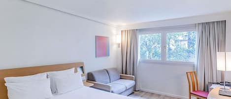 Habitación de hotel con cama, sofá, escritorio con silla, ventana con cortinas y una obra de arte colgada en la pared.