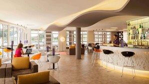 A modern bar area with a bar counter, seating with stools, and a person working on a laptop. - Novotel Setubal (Setubal)