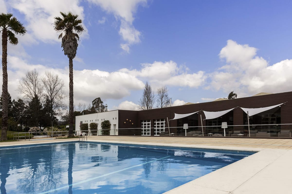 Une piscine, un ciel bleu azur parsemé de nuages, un bâtiment doté d’un toit plat et de grandes fenêtres, et des palmiers en arrière-plan.