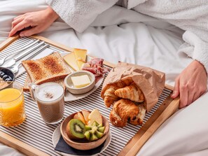 A breakfast tray with toast, cheese, ham, croissants, fruit, and juice on a bed.