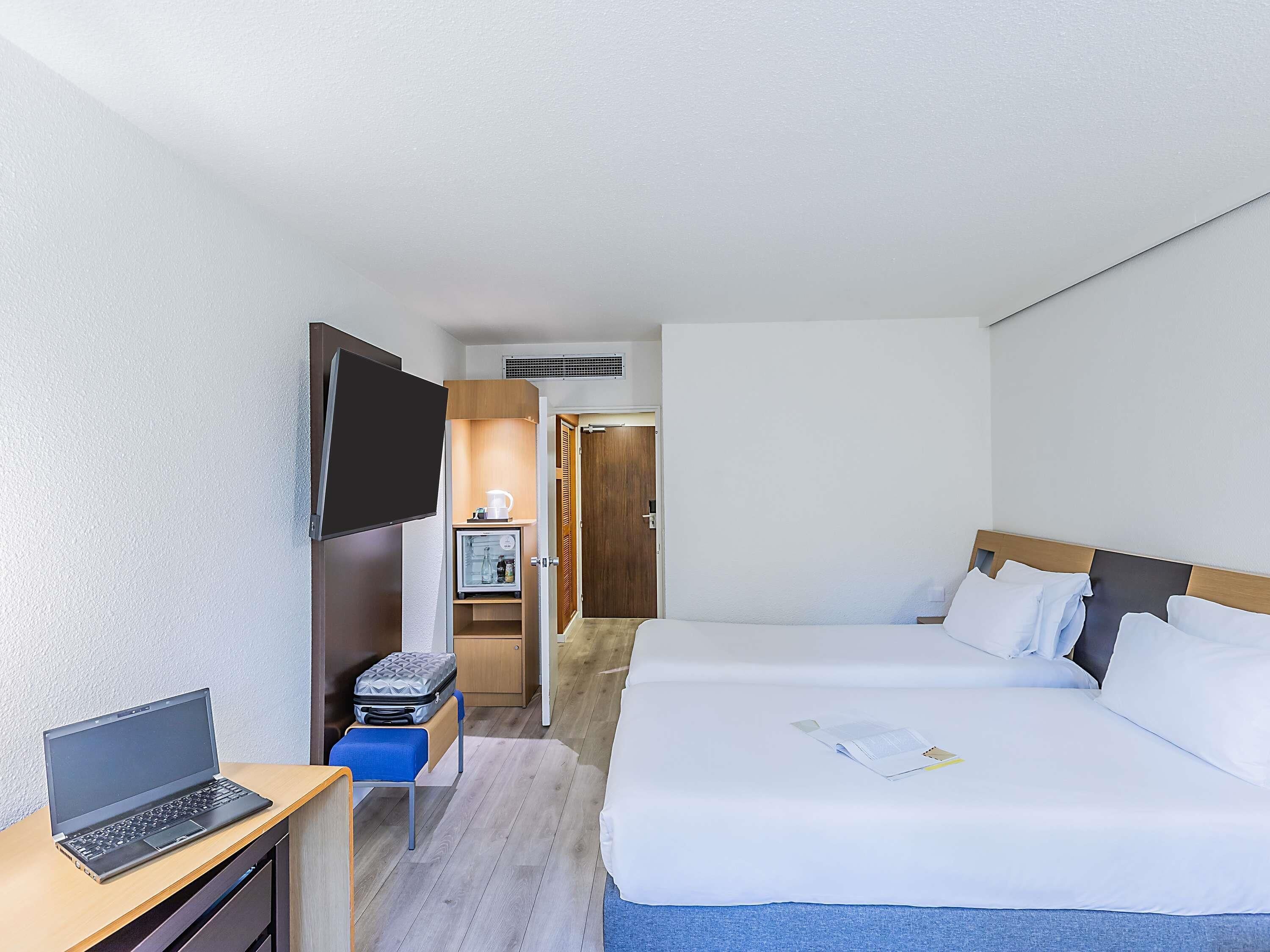 Hotellihuone, jossa on sänky, työpöytä kannettavalla tietokoneella, minijääkaappi ja seinälle asennettu televisio.