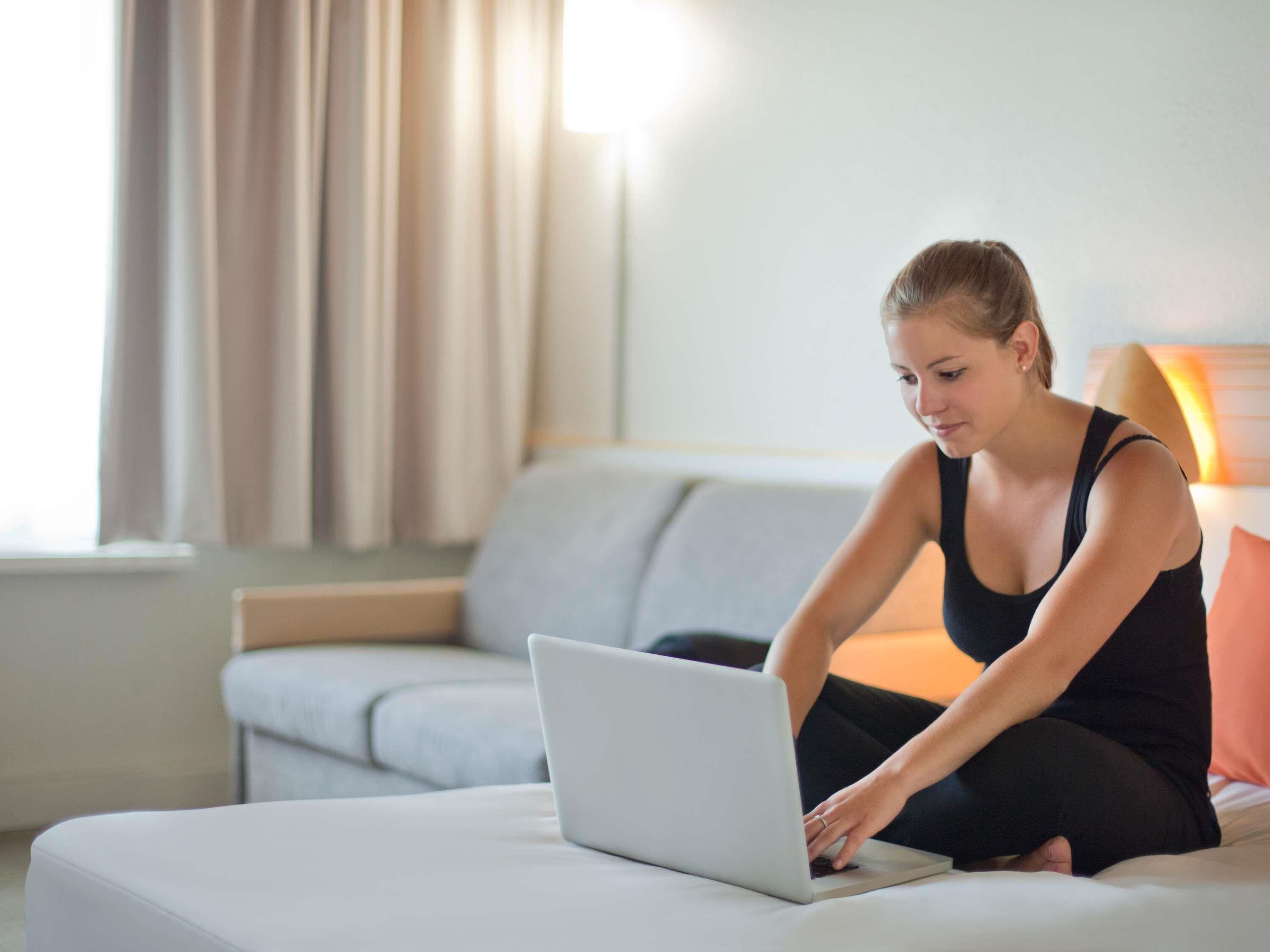 Una persona sentada en un sofá usando una computadora portátil en una habitación con ventana y cortinas.