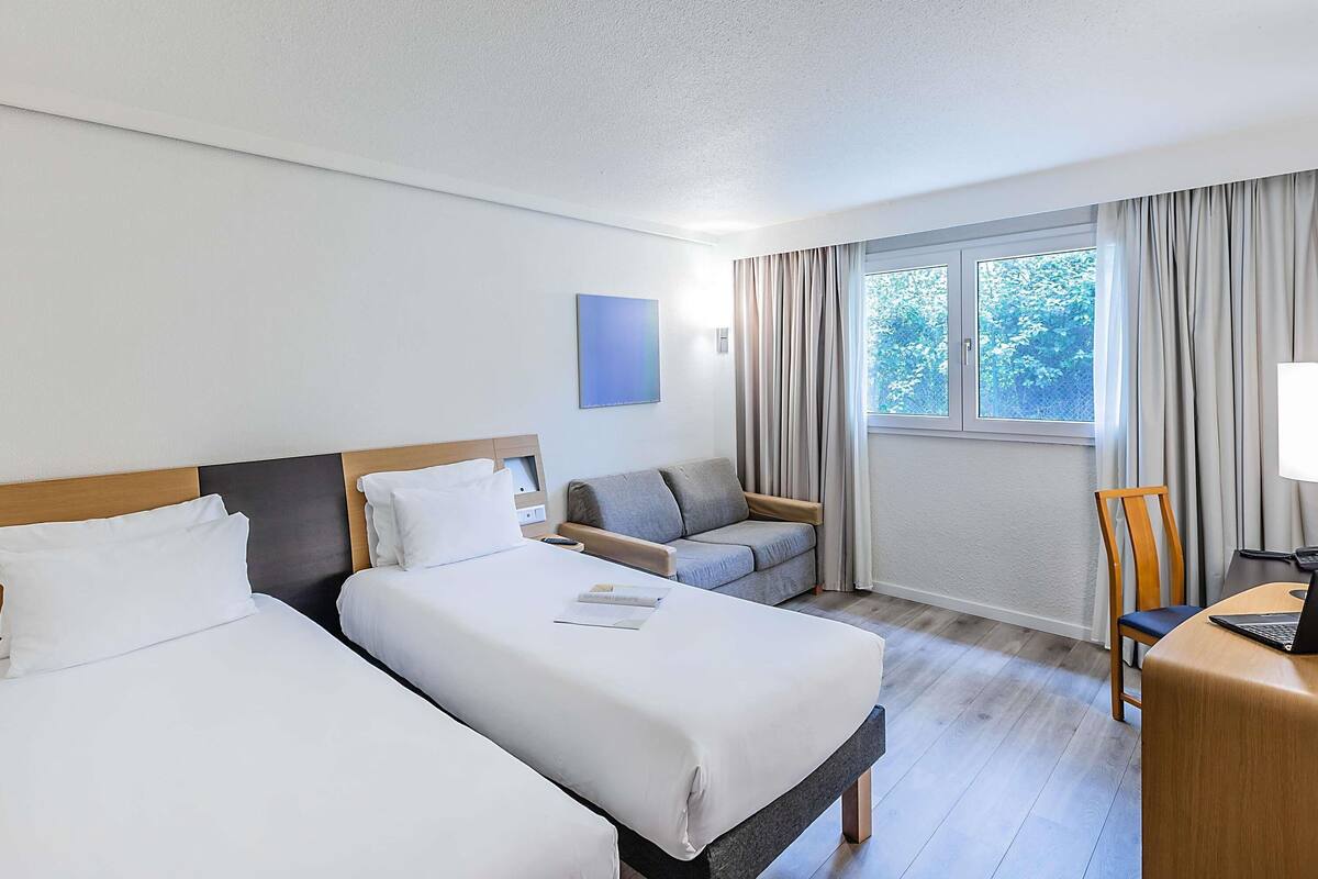 Habitación de hotel con dos camas, un escritorio, una silla, una ventana con cortinas y un cuadro en la pared.