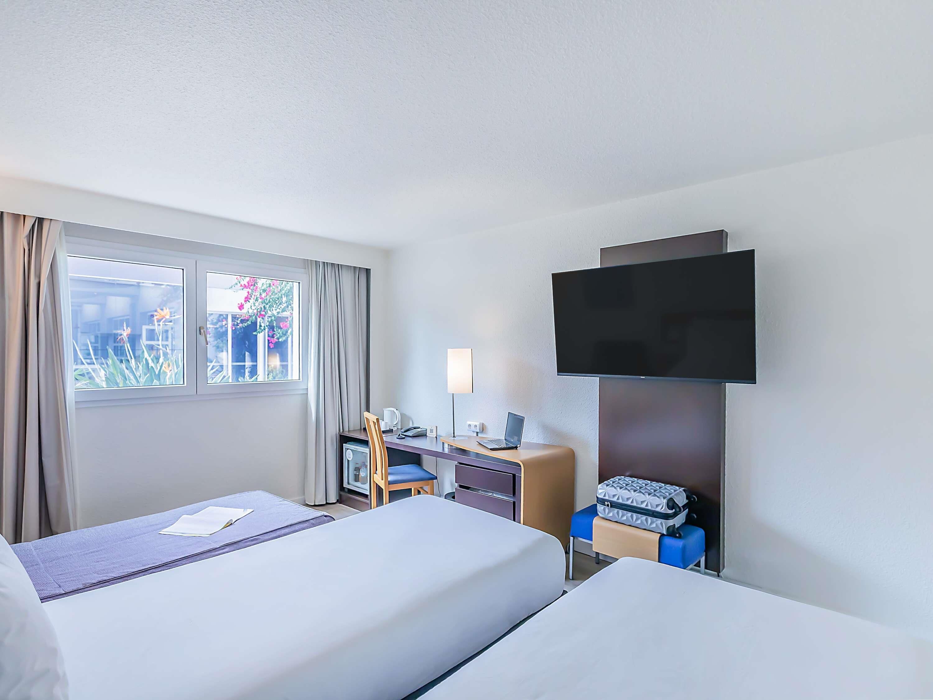 A hotel room with two beds, a desk with a computer, a TV mounted on the wall, and a window with curtains.