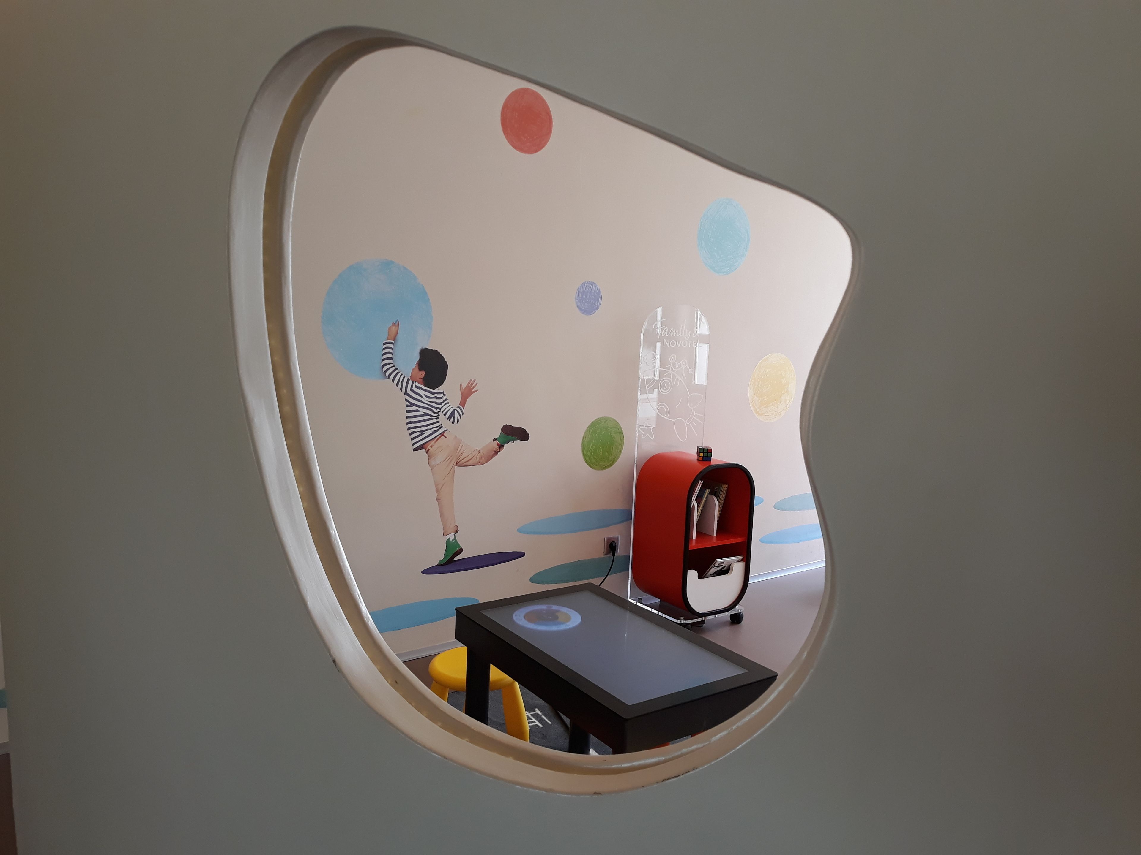 A child's artwork on a wall, a red bookshelf, and a small table with a yellow stool.
