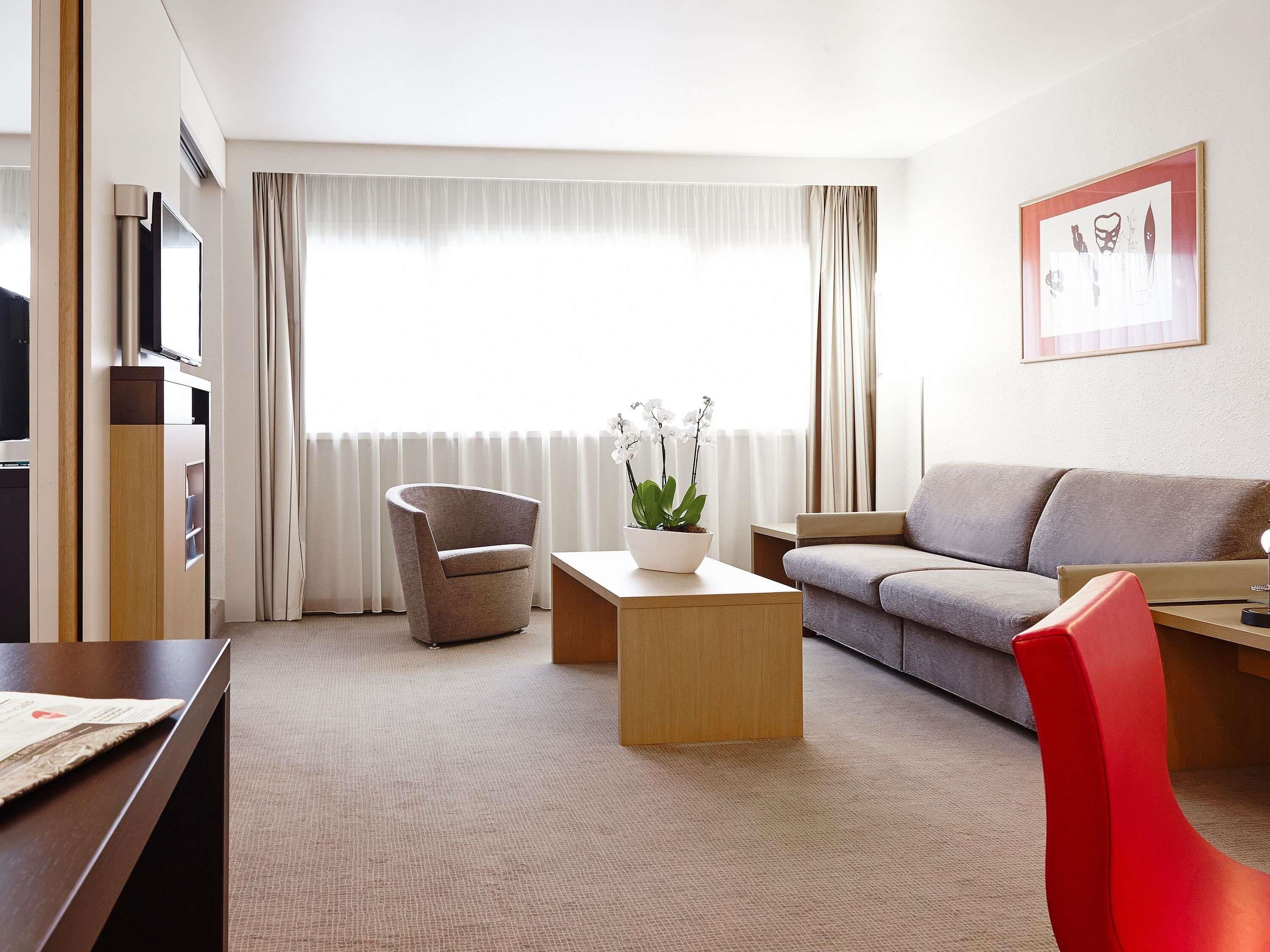 Ein modernes Hotelzimmer mit Sofa, Ohrensessel, Couchtisch und Fernseher.