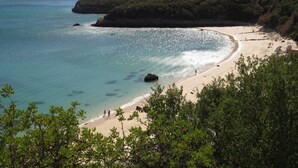 A coastal beach with clear turquoise waters, a sandy shore, and a backdrop of dense green hills. - Novotel Setubal (Setubal)