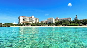 A beachfront hotel with clear turquoise water, a white building with a red roof, and a small white church in the background.