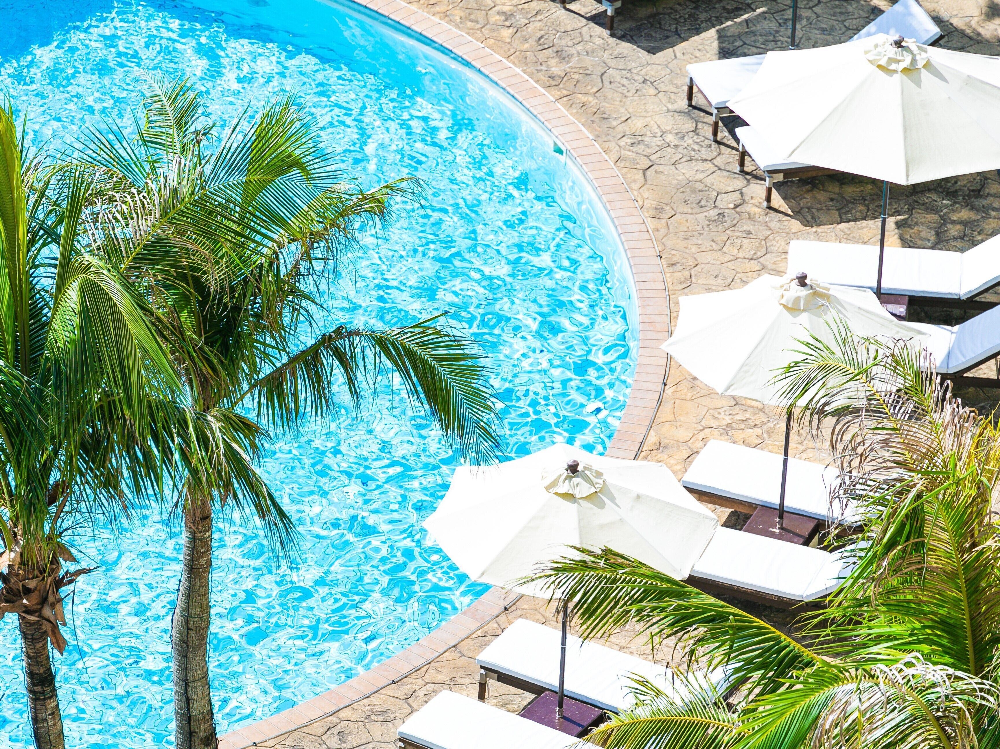 A circular pool with clear blue water, surrounded by stone tiles and palm trees. There are white lounge chairs and umbrellas around the pool.