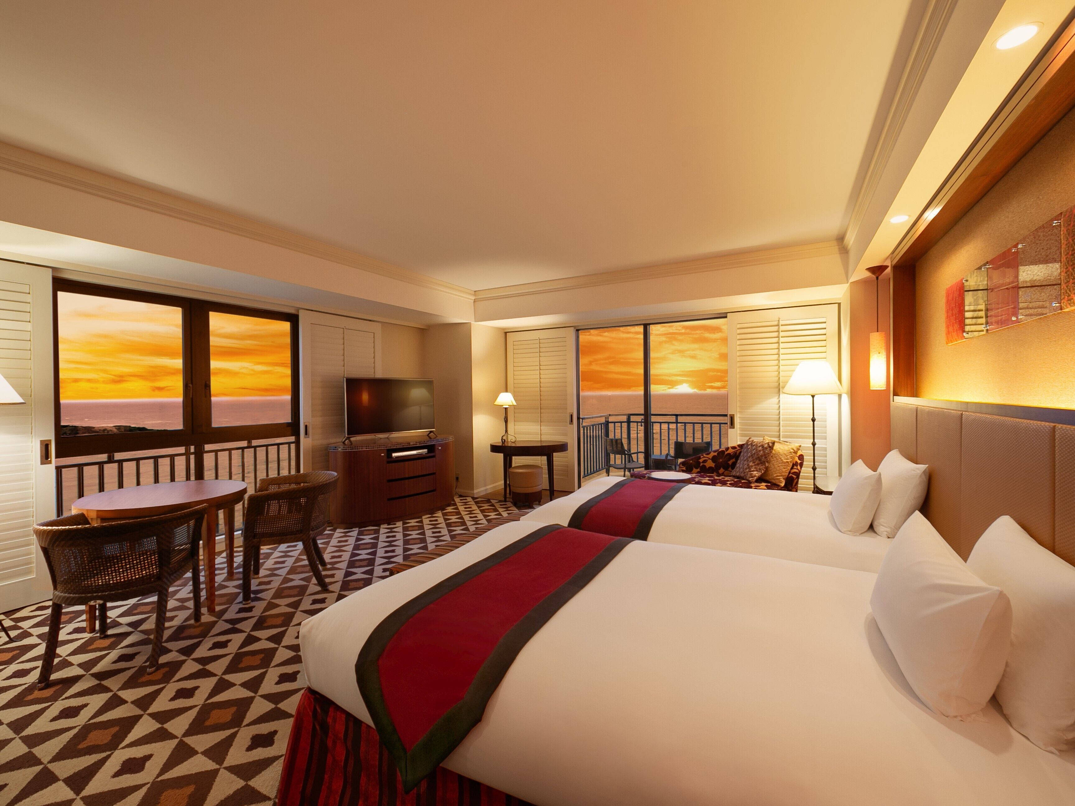 A hotel room with two beds, a TV, a desk with a chair, and a balcony with a sunset view.