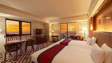 A hotel room with two beds, a TV, a desk with a chair, and a balcony with a sunset view.