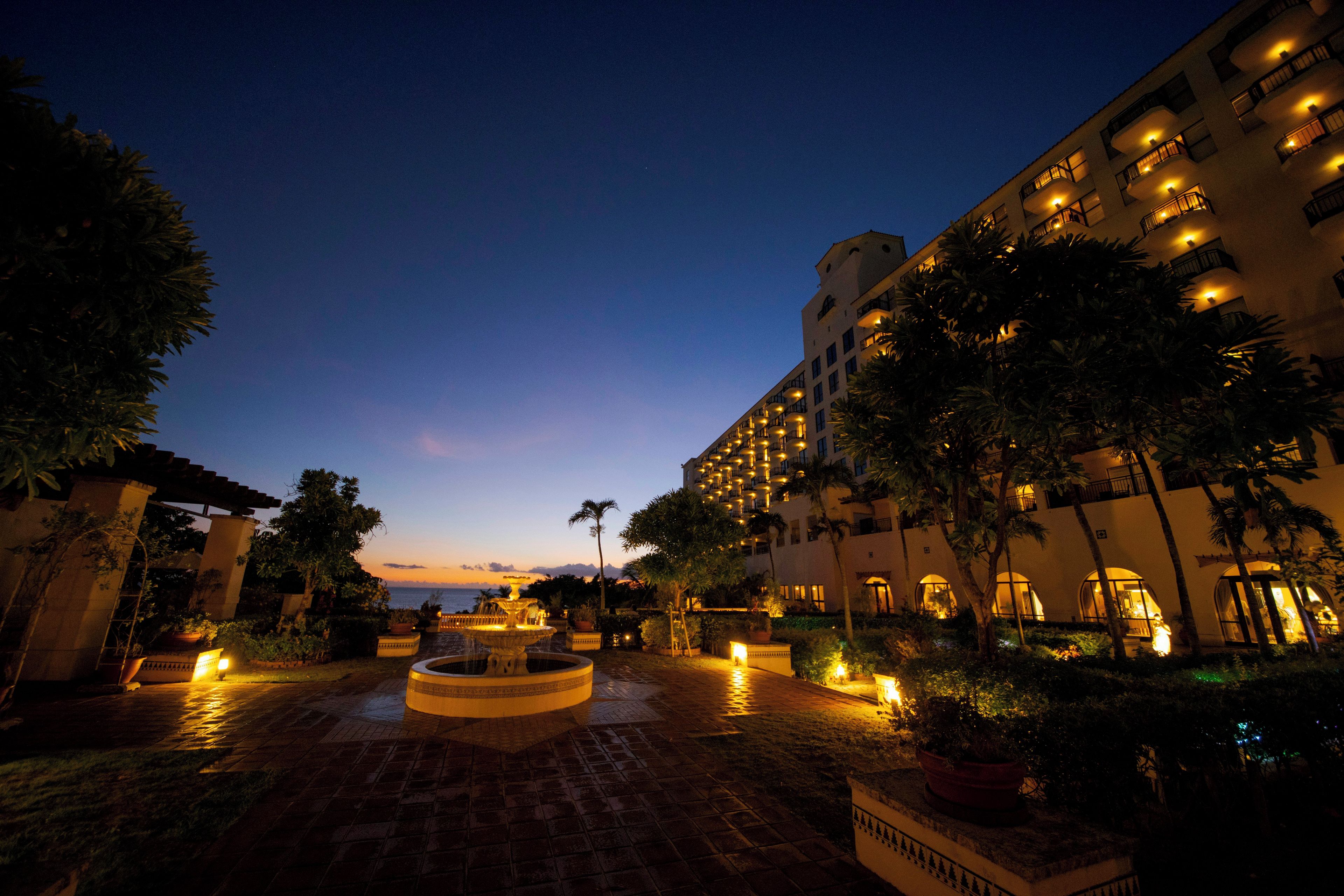 A courtyard with a central fountain, surrounded by palm trees and buildings with illuminated balconies.