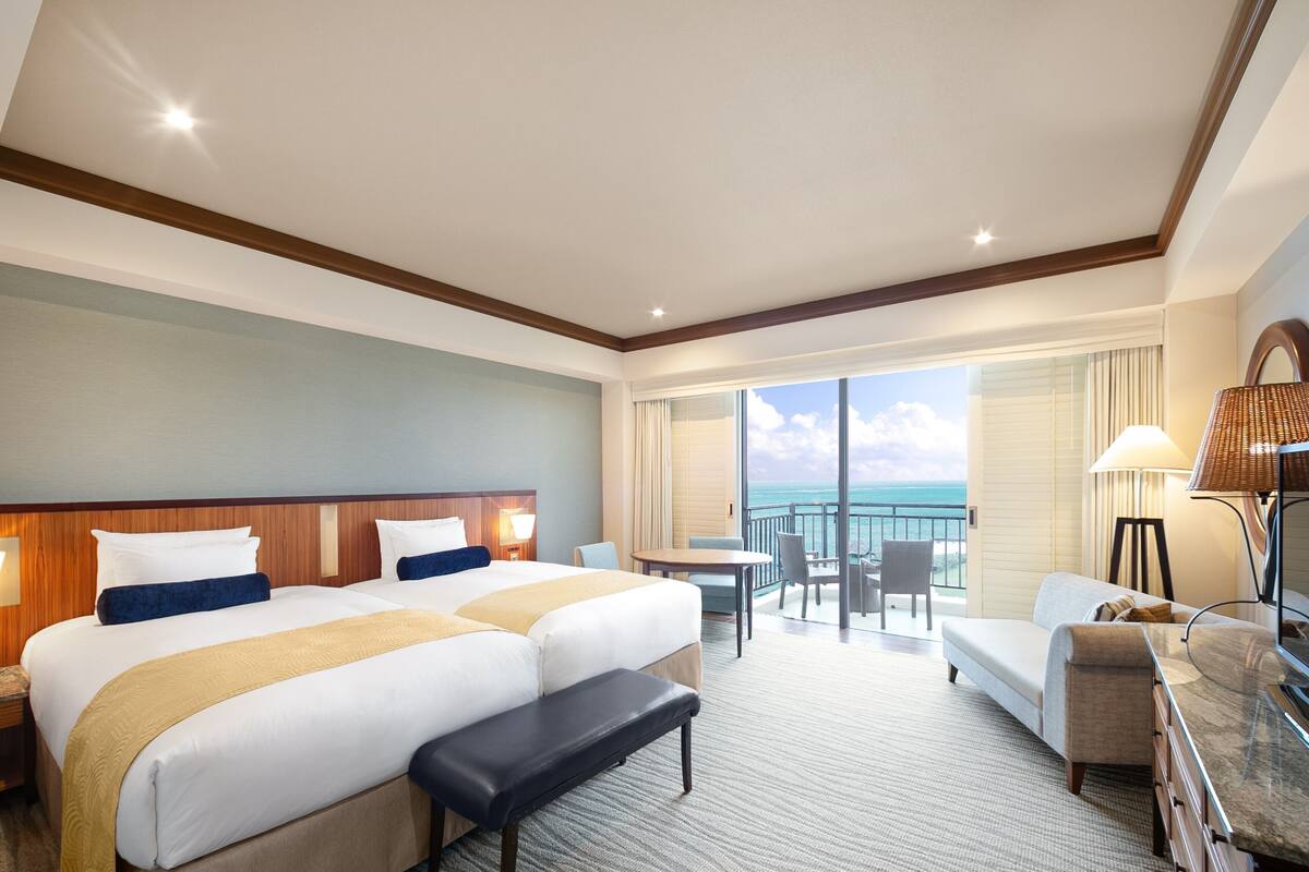 A hotel room with two beds, a desk, a chair, a sofa, a bench, and a view of the ocean.