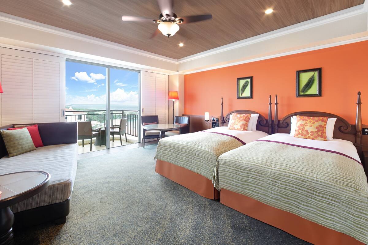 A hotel room with two beds, a balcony with a view, a ceiling fan, and a desk with a chair.