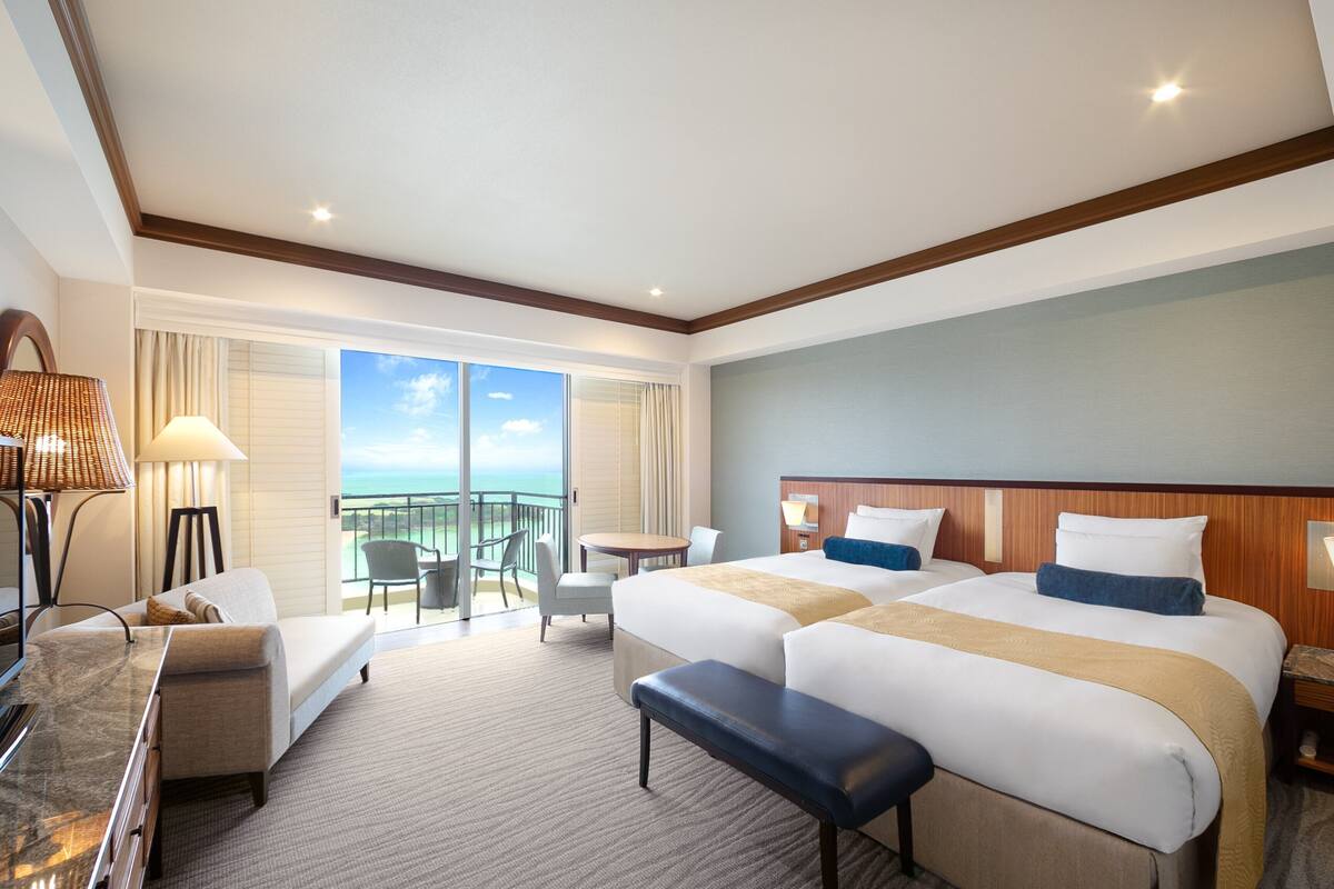 A hotel room with two beds, a desk, a chair, a bench, and a view of the ocean.