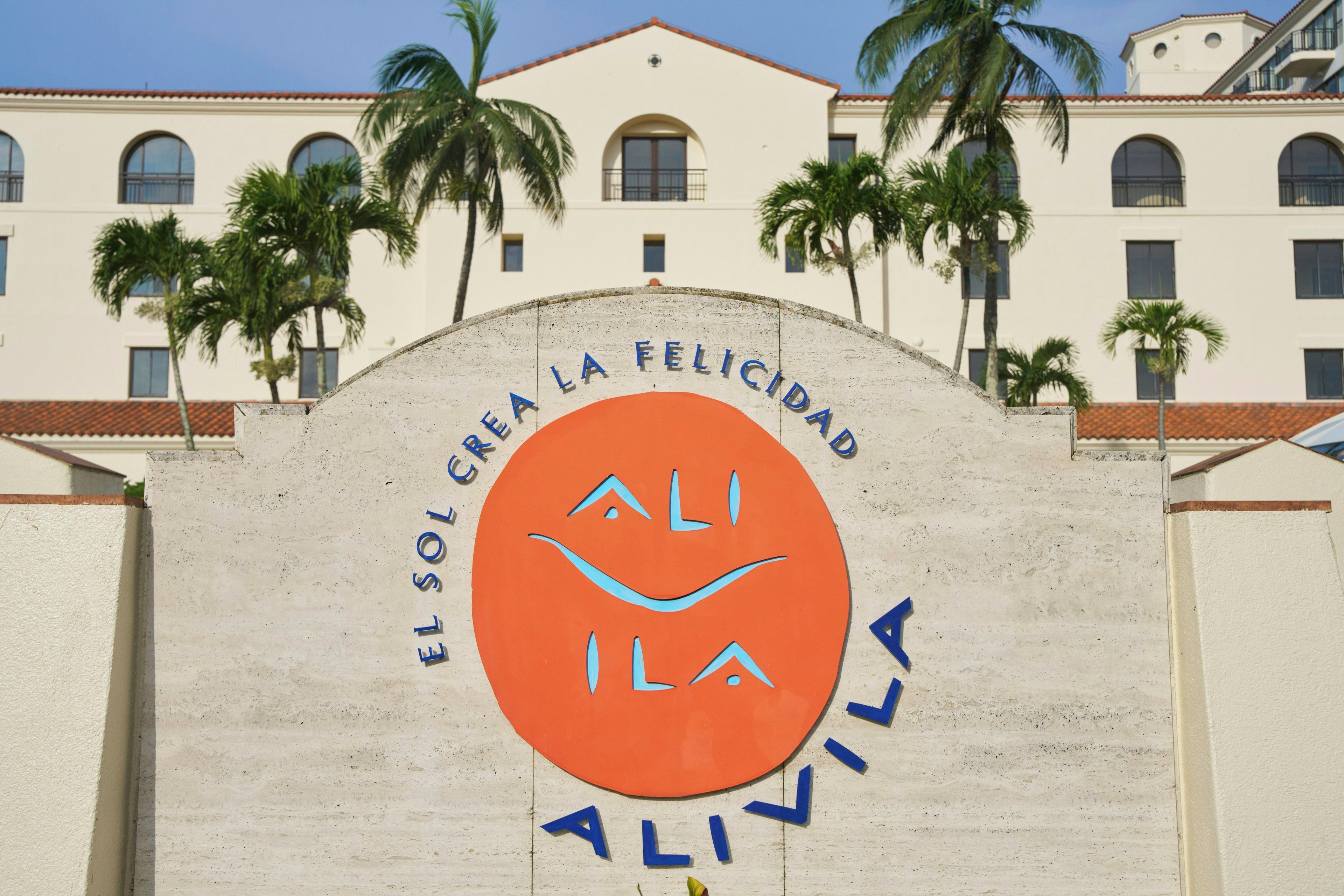A building with a sign that reads 'EL SOL CREA LA FELICIDAD' and 'ALI ILA ALIVILA' with palm trees in the background.