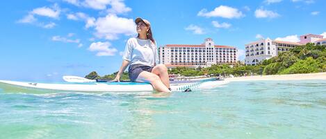 Una persona sentada sobre una tabla de surf en agua cristalina, con un hotel y una playa al fondo.