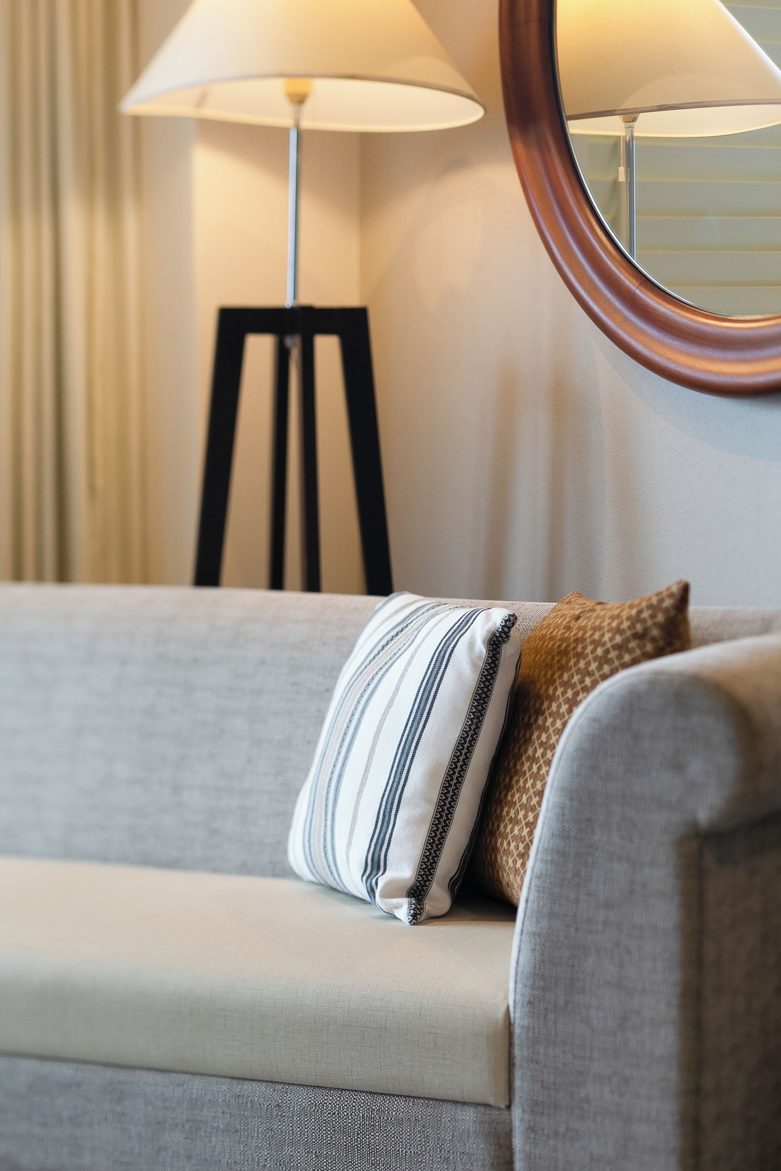A sofa with a striped pillow and a houndstooth pillow, a floor lamp, and a round mirror on the wall.