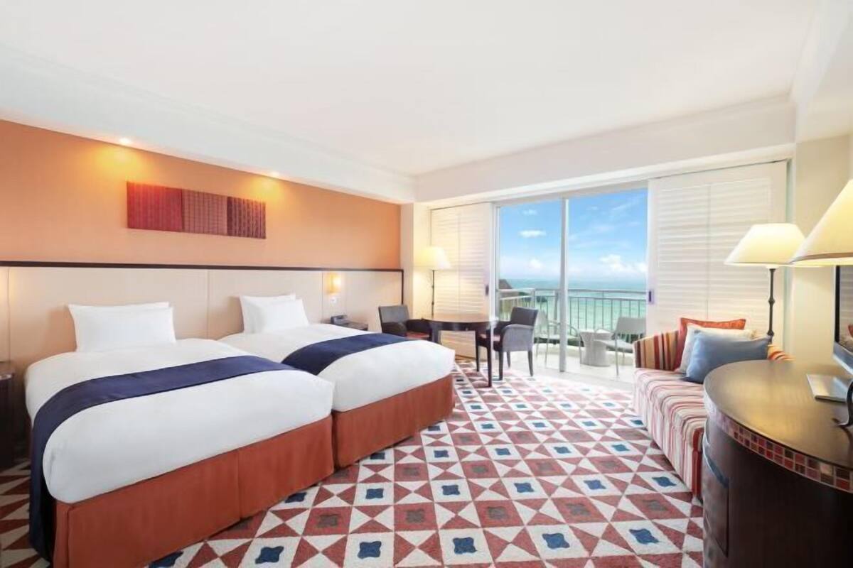 A hotel room with two beds, a desk, a sofa, and a balcony with a view.