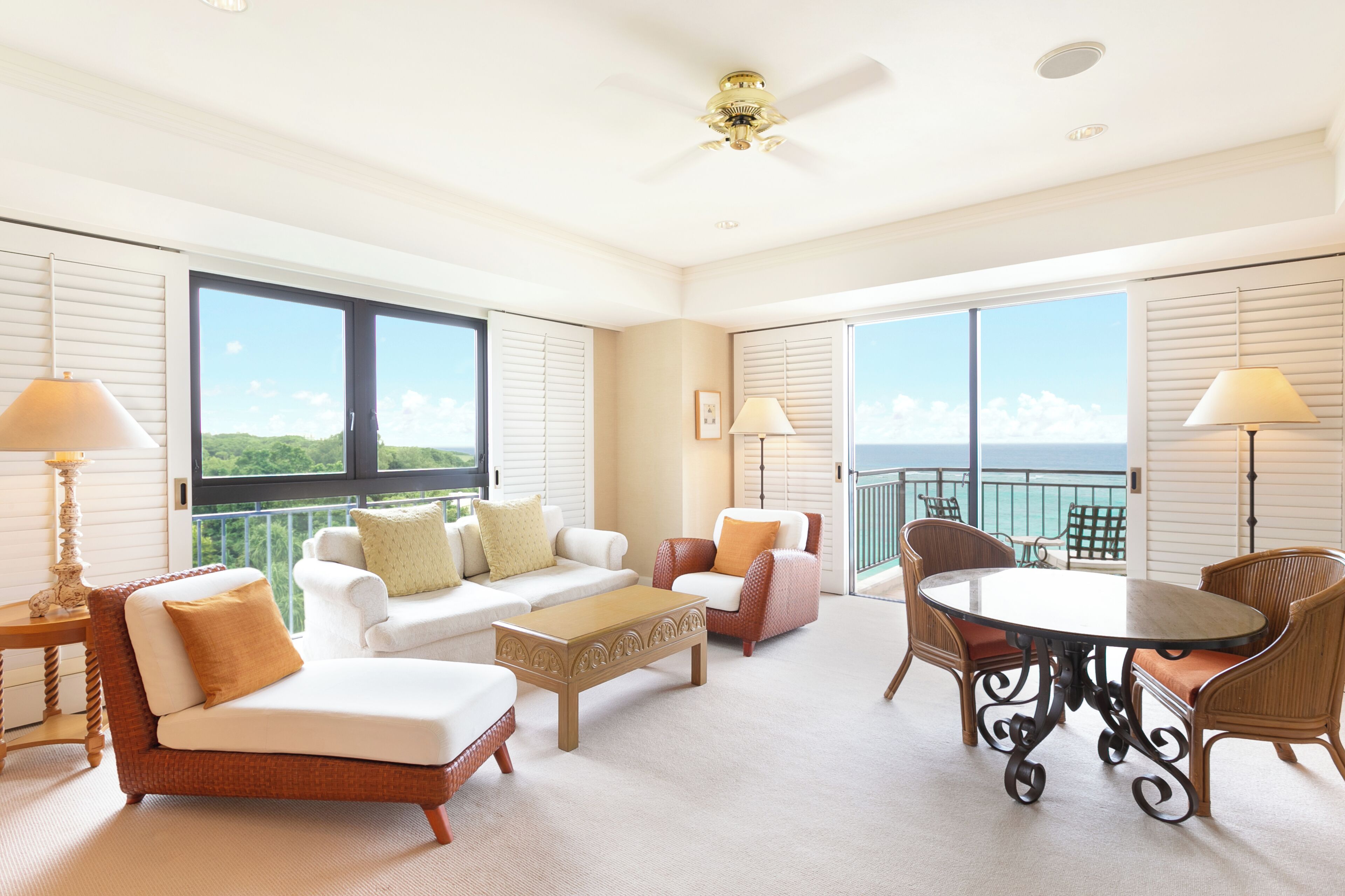 A spacious balcony with a white ceiling fan, a white sofa with cushions, a wooden coffee table, wicker chairs, and a round table with chairs.