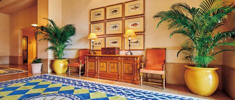 A lobby with a patterned carpet, a wooden reception desk, potted plants, and framed fish pictures on the wall.