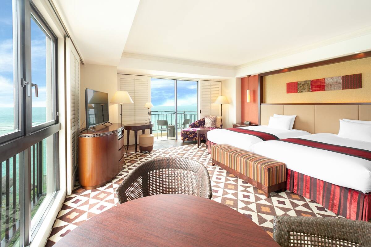 A hotel room with a large bed, a desk, a TV, a balcony with a view of the ocean, and a patterned floor.
