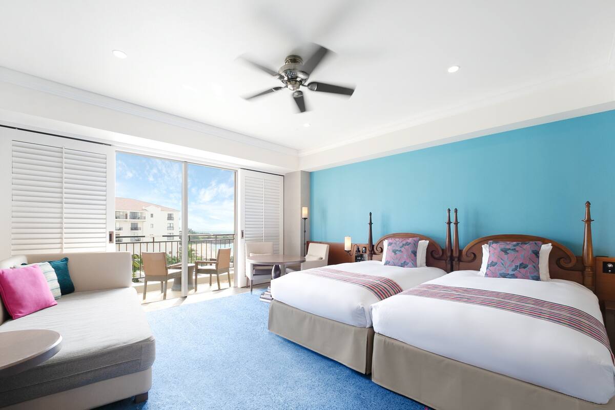 A hotel room with two beds, a sofa, a balcony with a table and chairs, and a ceiling fan.