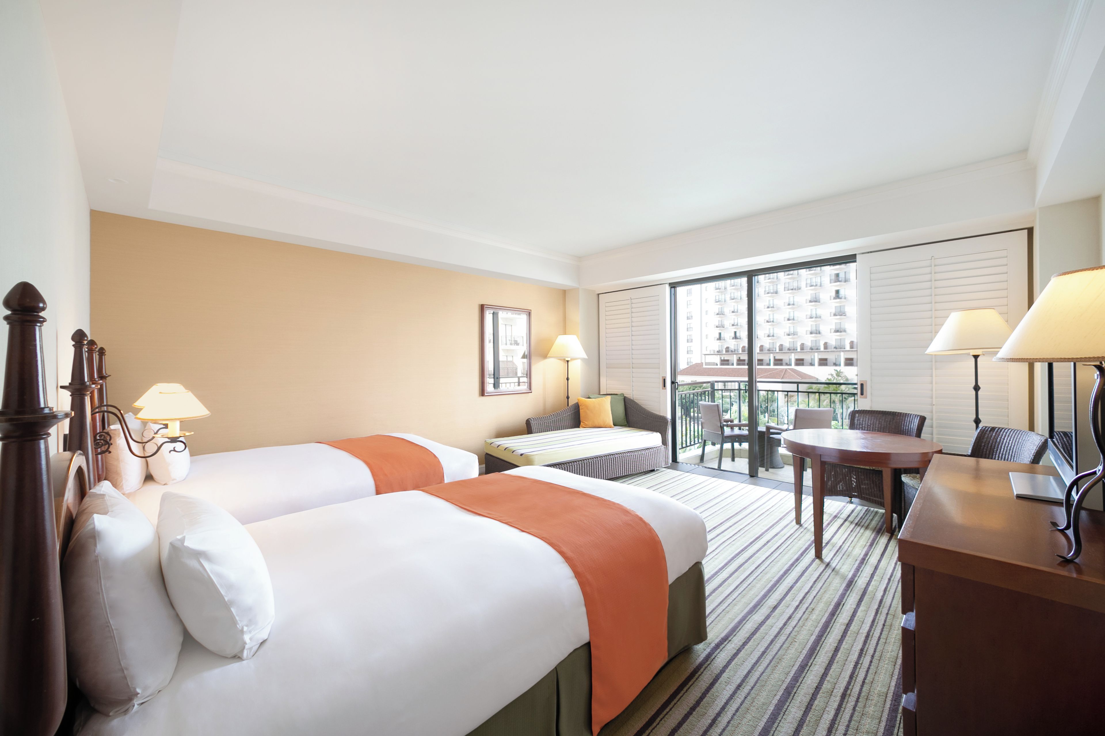A hotel room with two beds, a desk, a chair, and a balcony with a view of buildings.