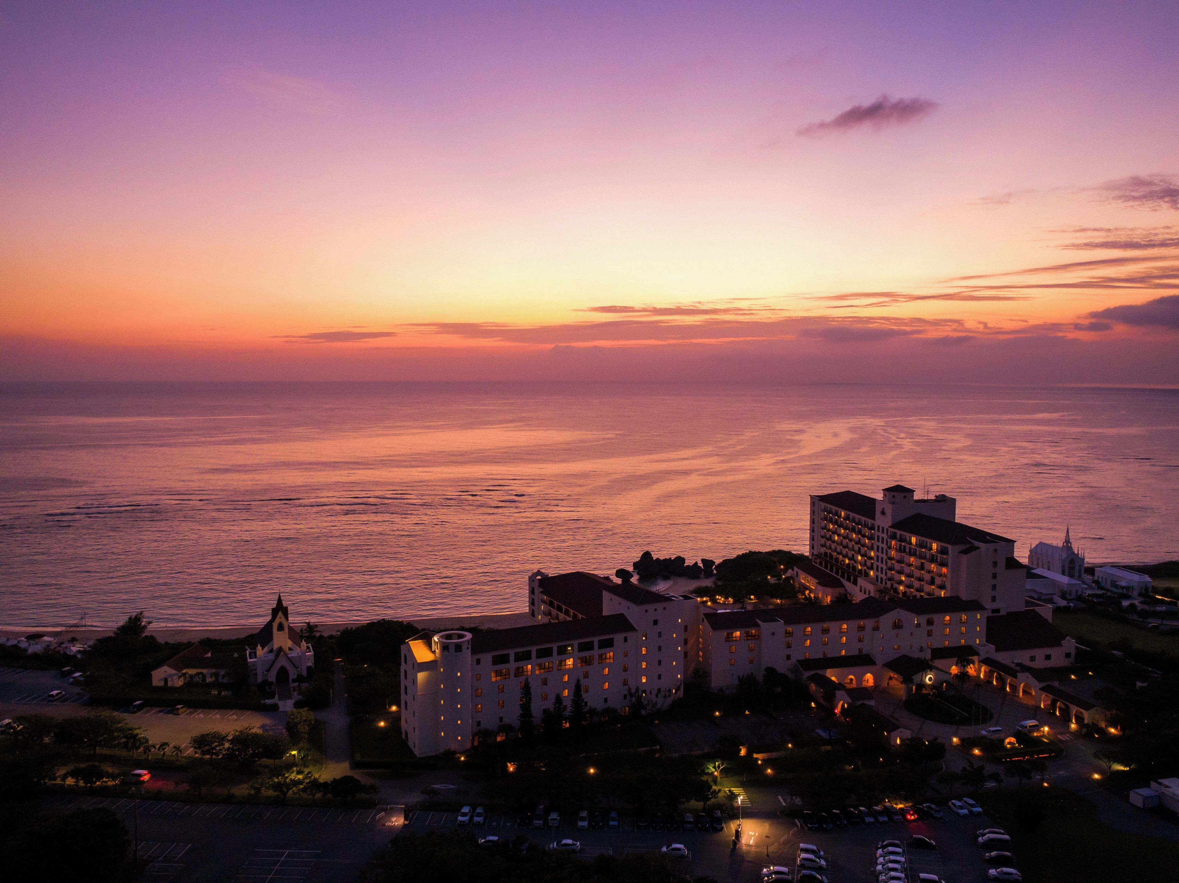 Un hotel costero con varias edificaciones, un estacionamiento y vista al mar al atardecer.