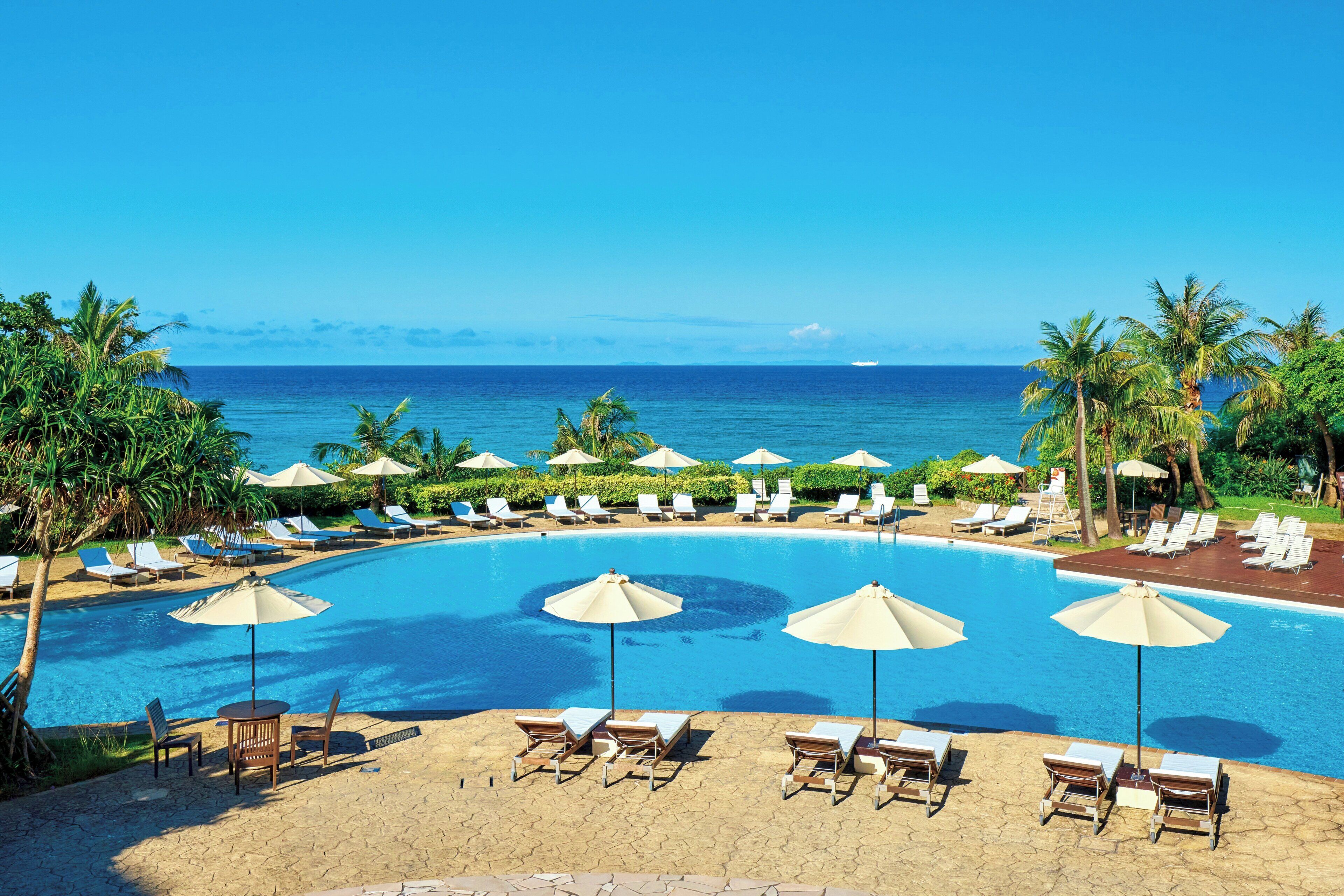 Zona de piscina con sillones reclinables y sombrillas, con vistas a un océano azul y despejado.