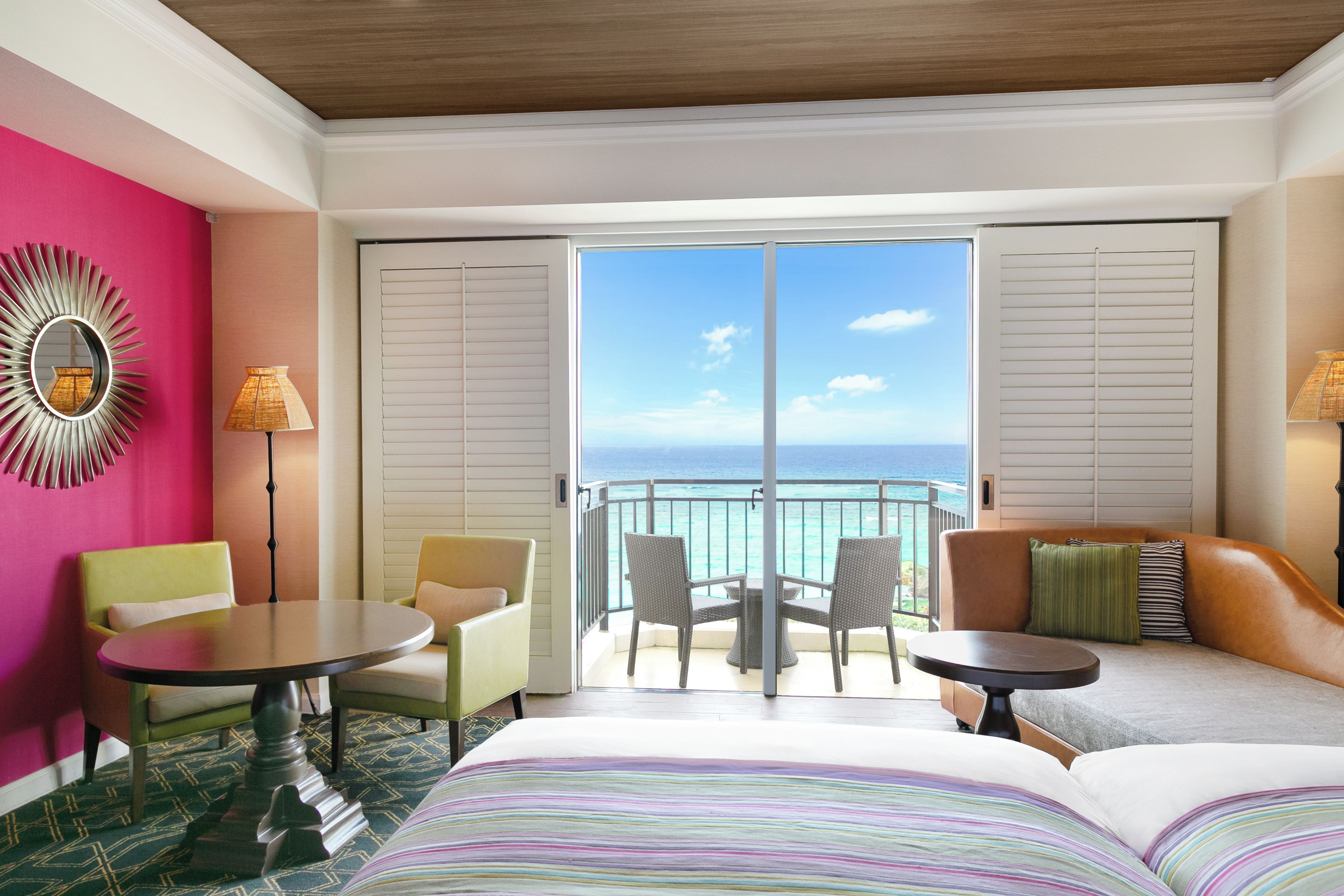 Habitación de hotel con balcón, cama con ropa de cama a rayas, sillón verde, mesa redonda y vista al océano.