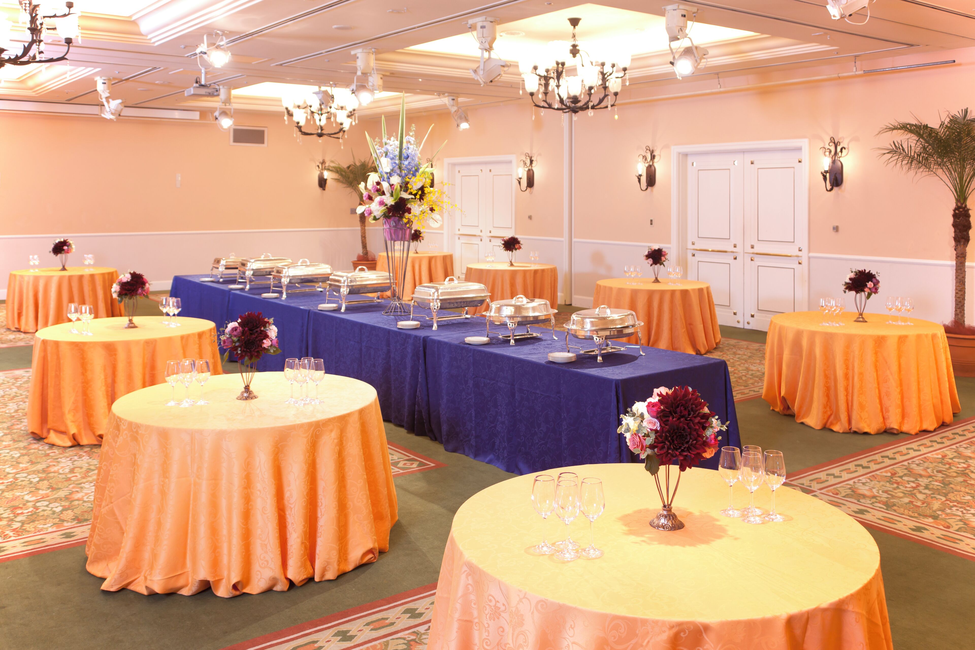 Un salón de banquetes con mesas redondas cubiertas con manteles naranja, una mesa central con mantel azul y centros de mesa florales.
