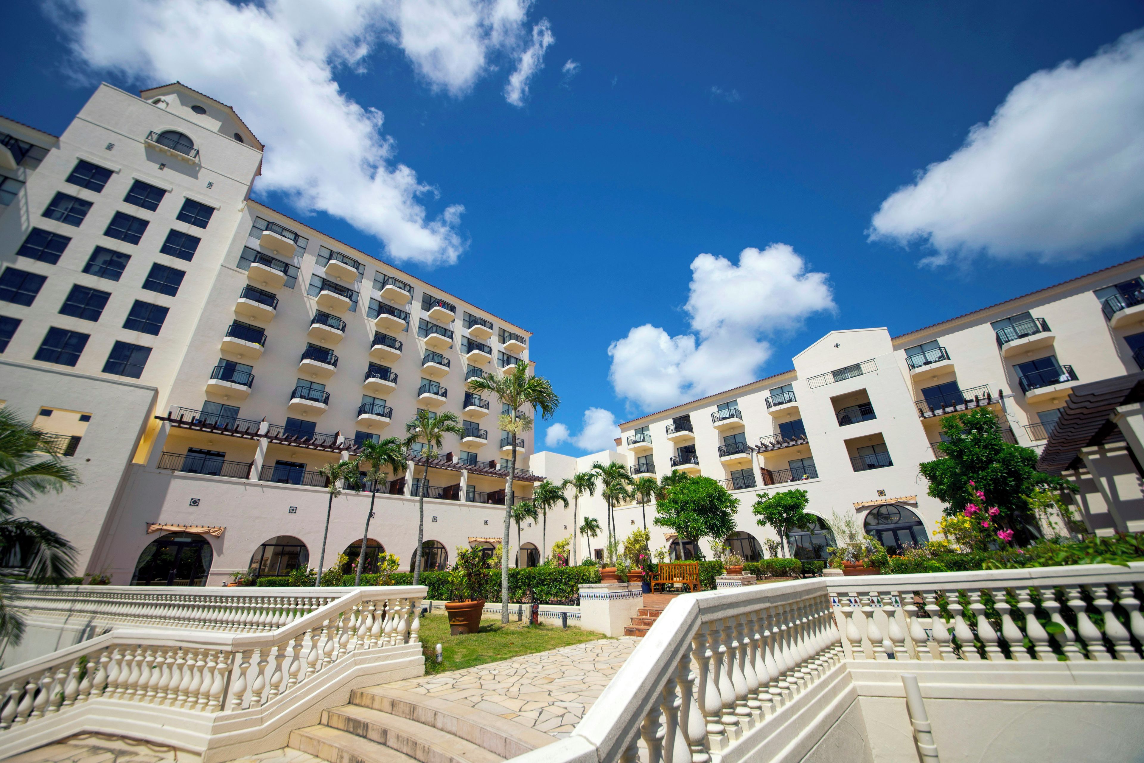 A multi-story hotel with balconies, a central courtyard with palm trees, and a staircase leading to the entrance.