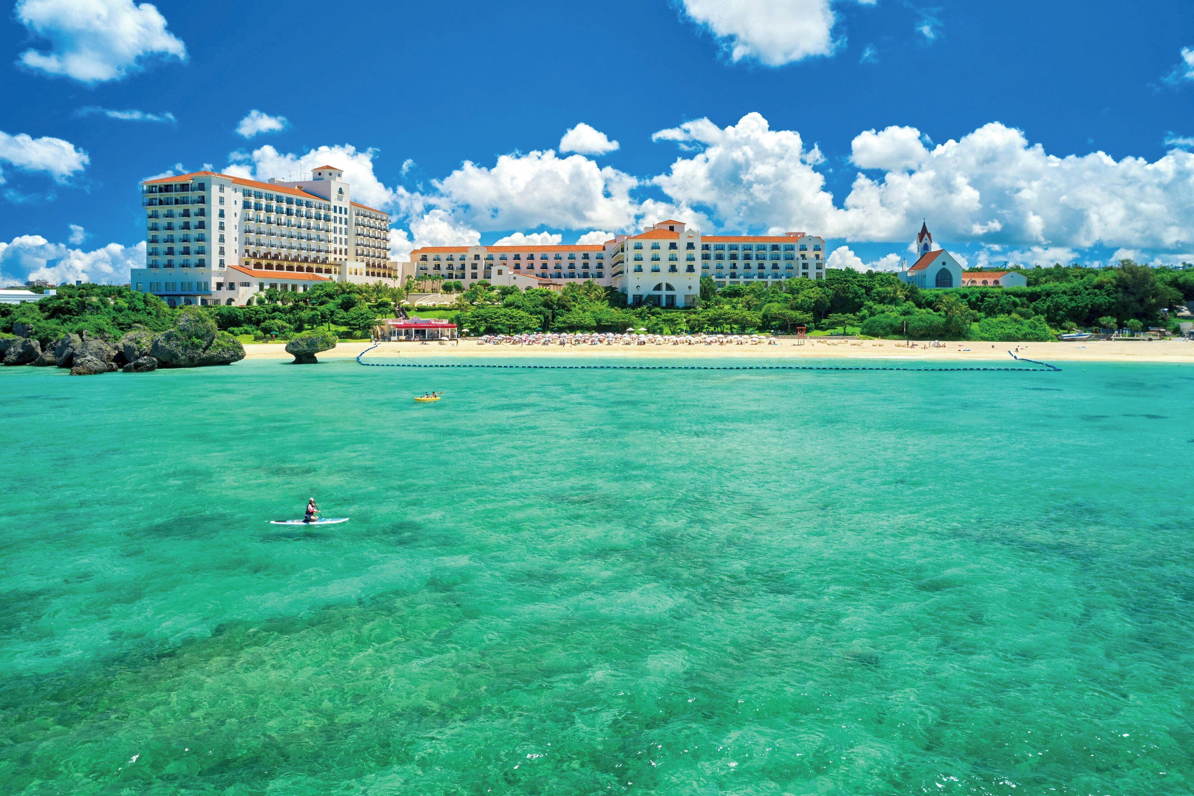 A beachfront hotel with multiple buildings, a clear blue sky, and a person kayaking in the water.