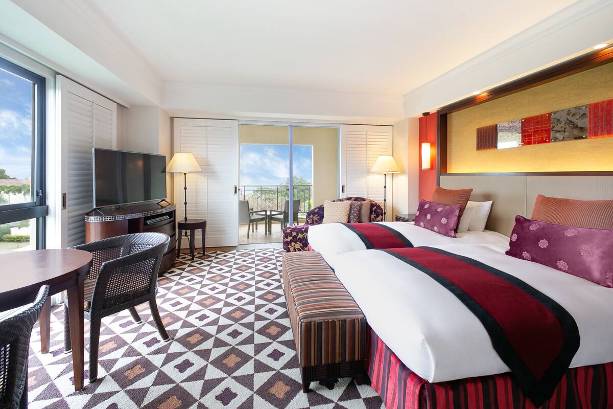 A hotel room with a large bed, a TV, a dining table, and a balcony with a view.