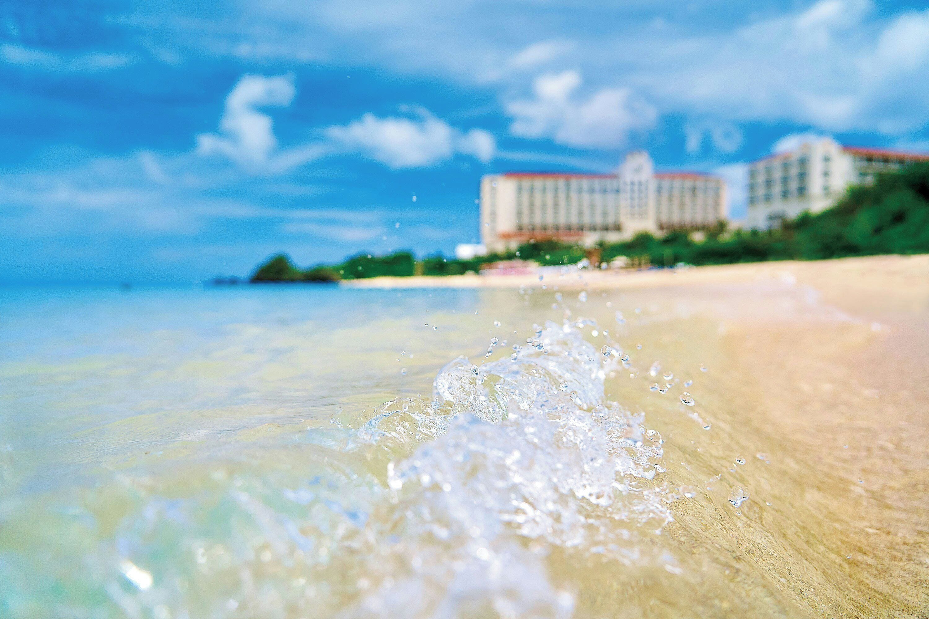 A beach with clear water, a sandy shore, and a building in the background.