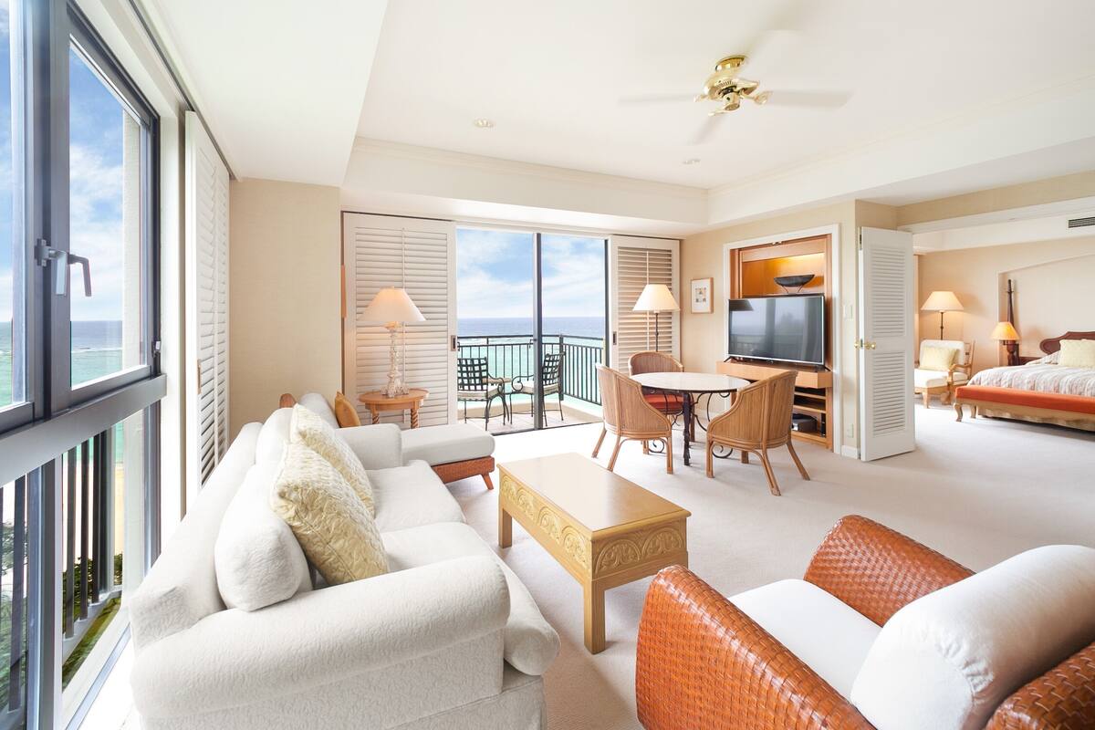 A spacious living room with a balcony, a dining area, and a bedroom with a TV.