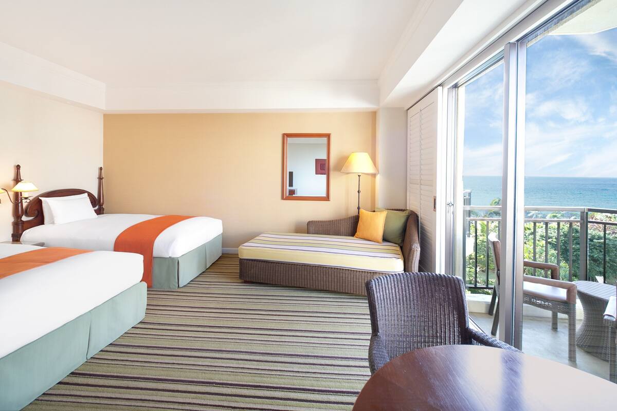 A hotel room with two beds, a balcony with a view of the ocean, a large window, and a chair.