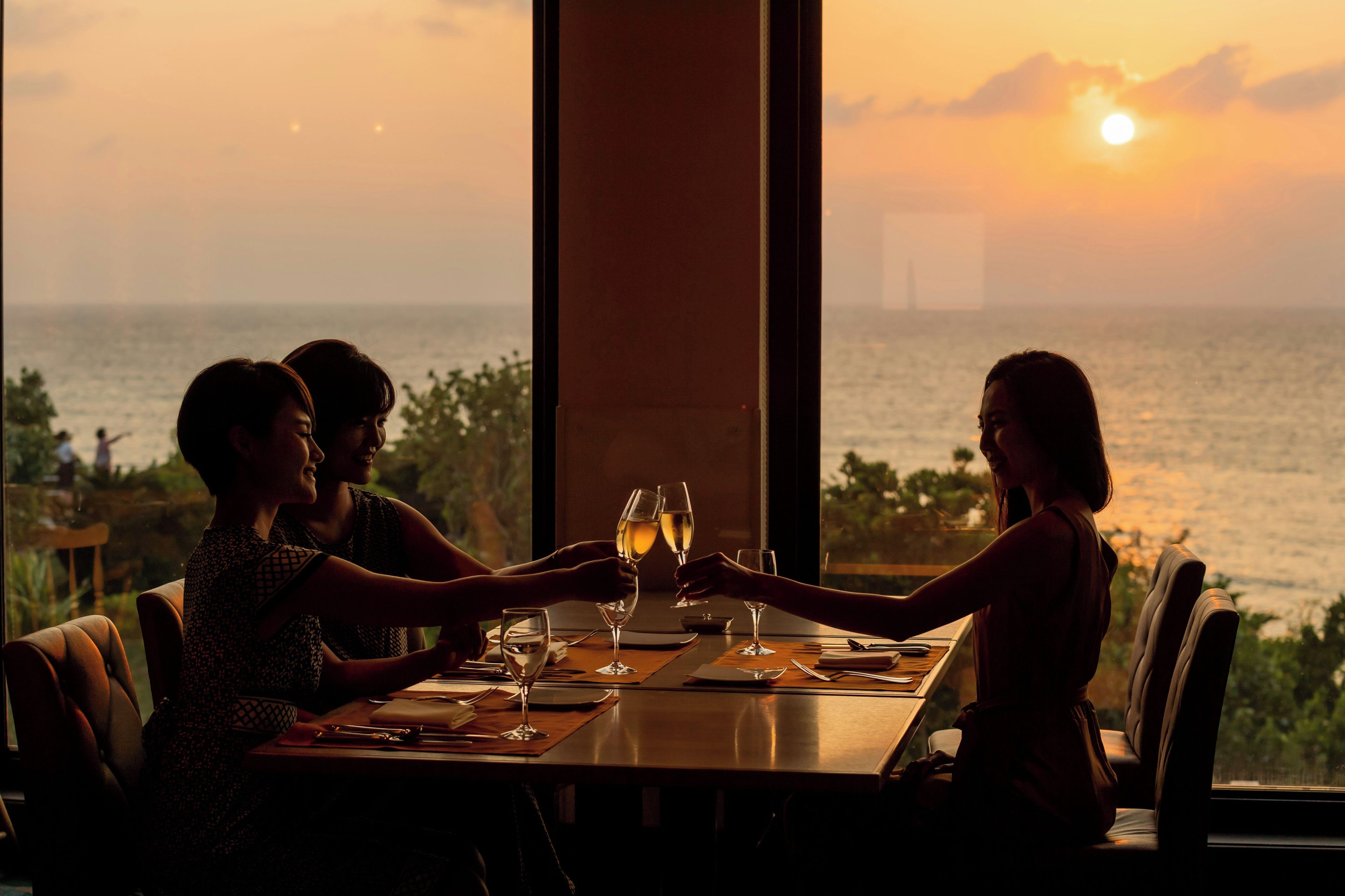 A table set for three people with glasses and cutlery, overlooking a body of water at sunset.