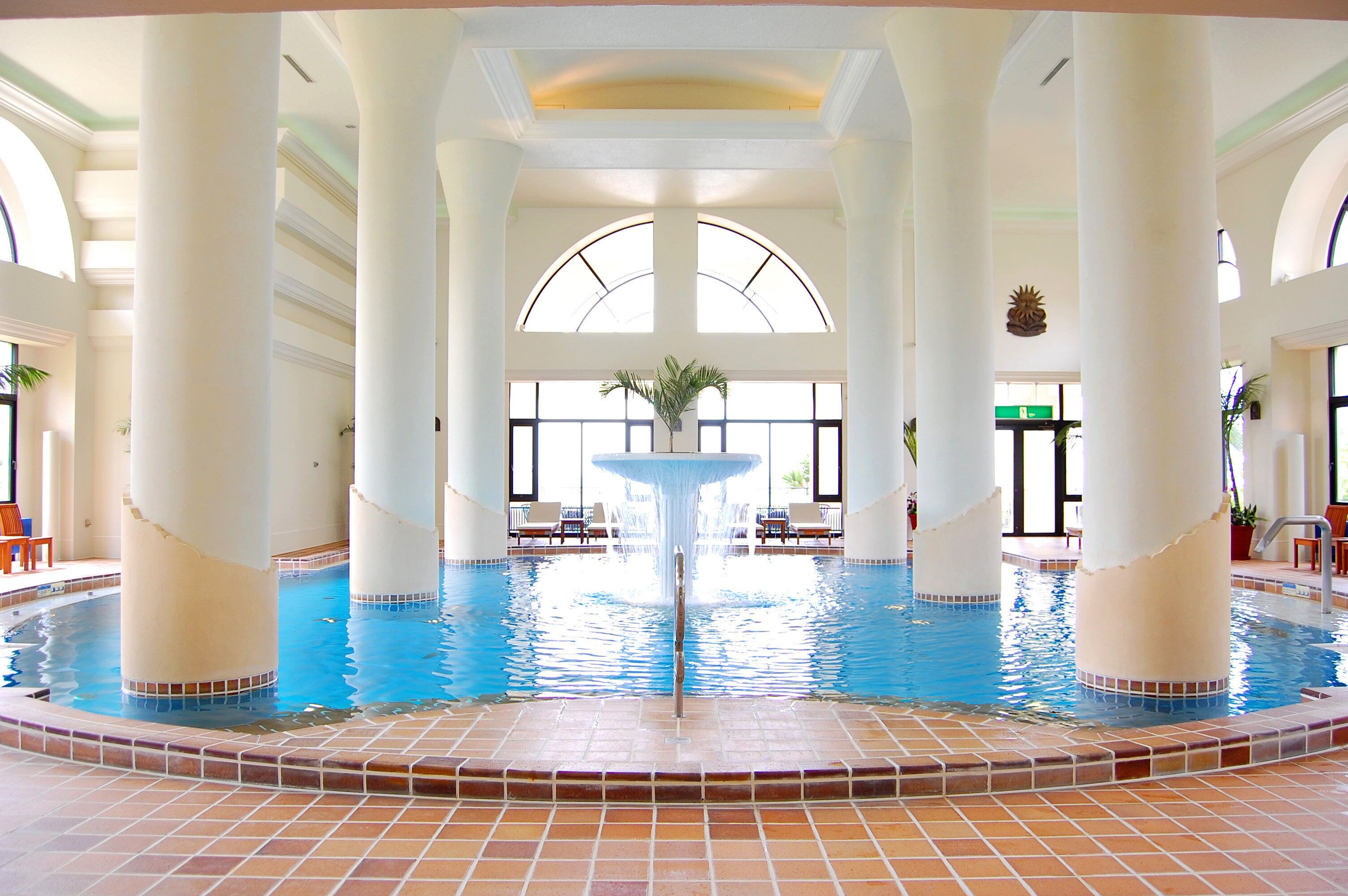 Una piscina interior con una fuente central, rodeada de columnas y sillas, con ventanas grandes y suelo de baldosas.