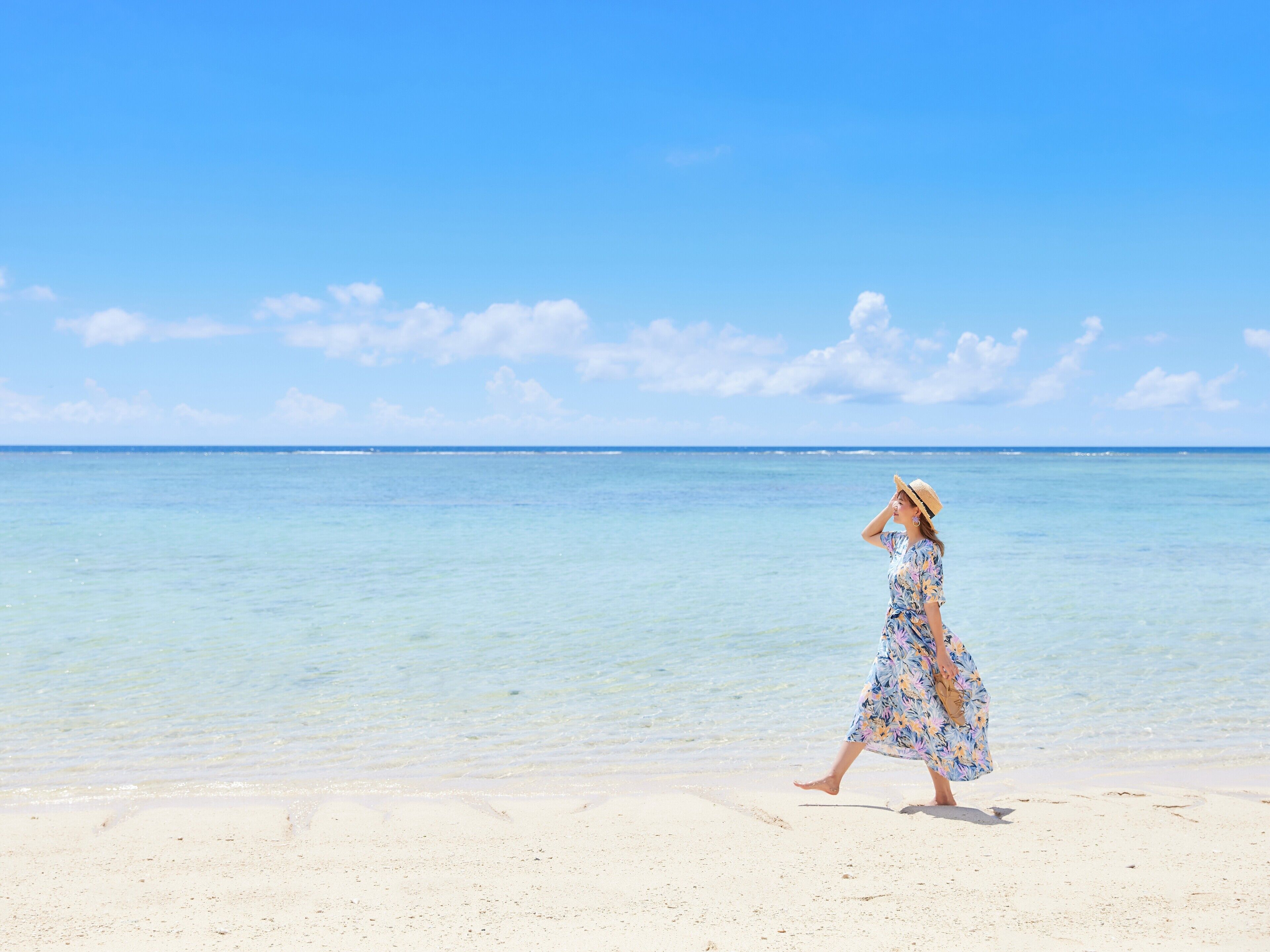 A woman walking on a sandy beach with clear blue water and a bright sky.