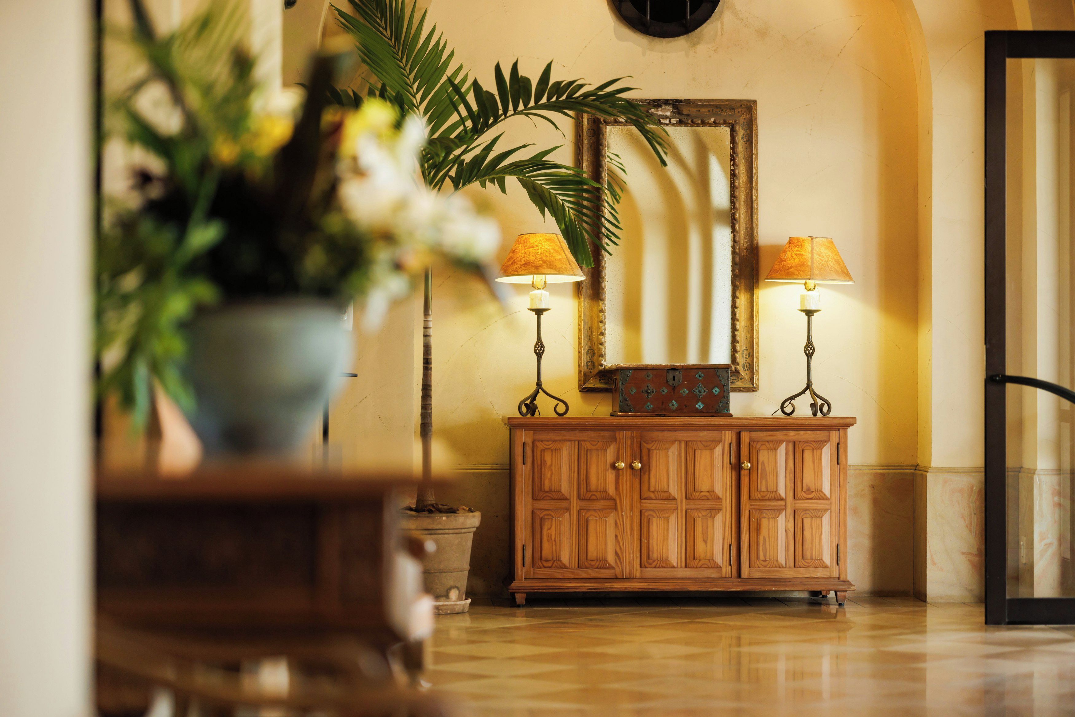 A wooden cabinet with two lamps, a mirror, and a potted plant in a lobby area.