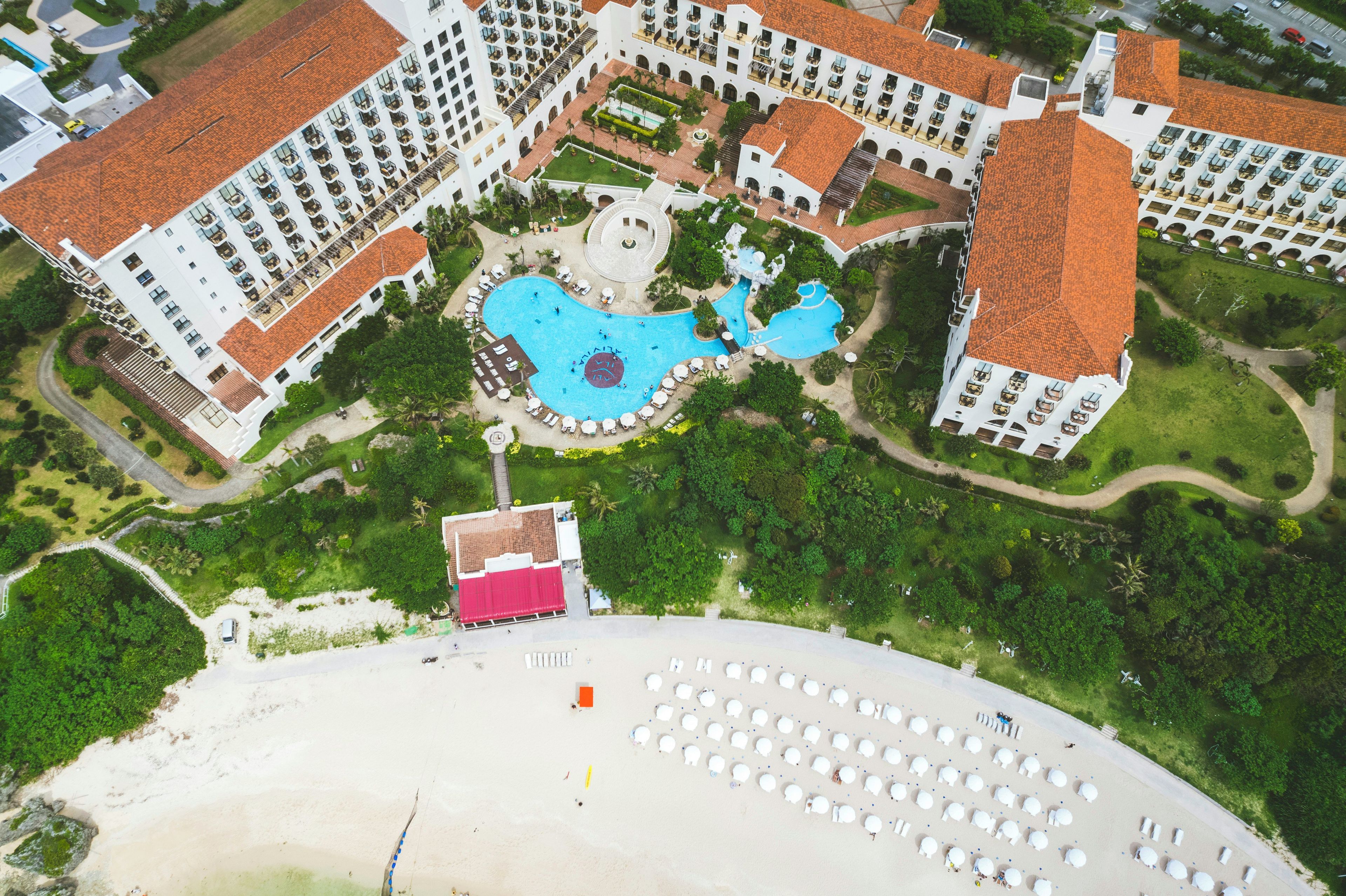 An aerial view of a resort with multiple buildings, a central swimming pool, and a beach area with sun loungers.