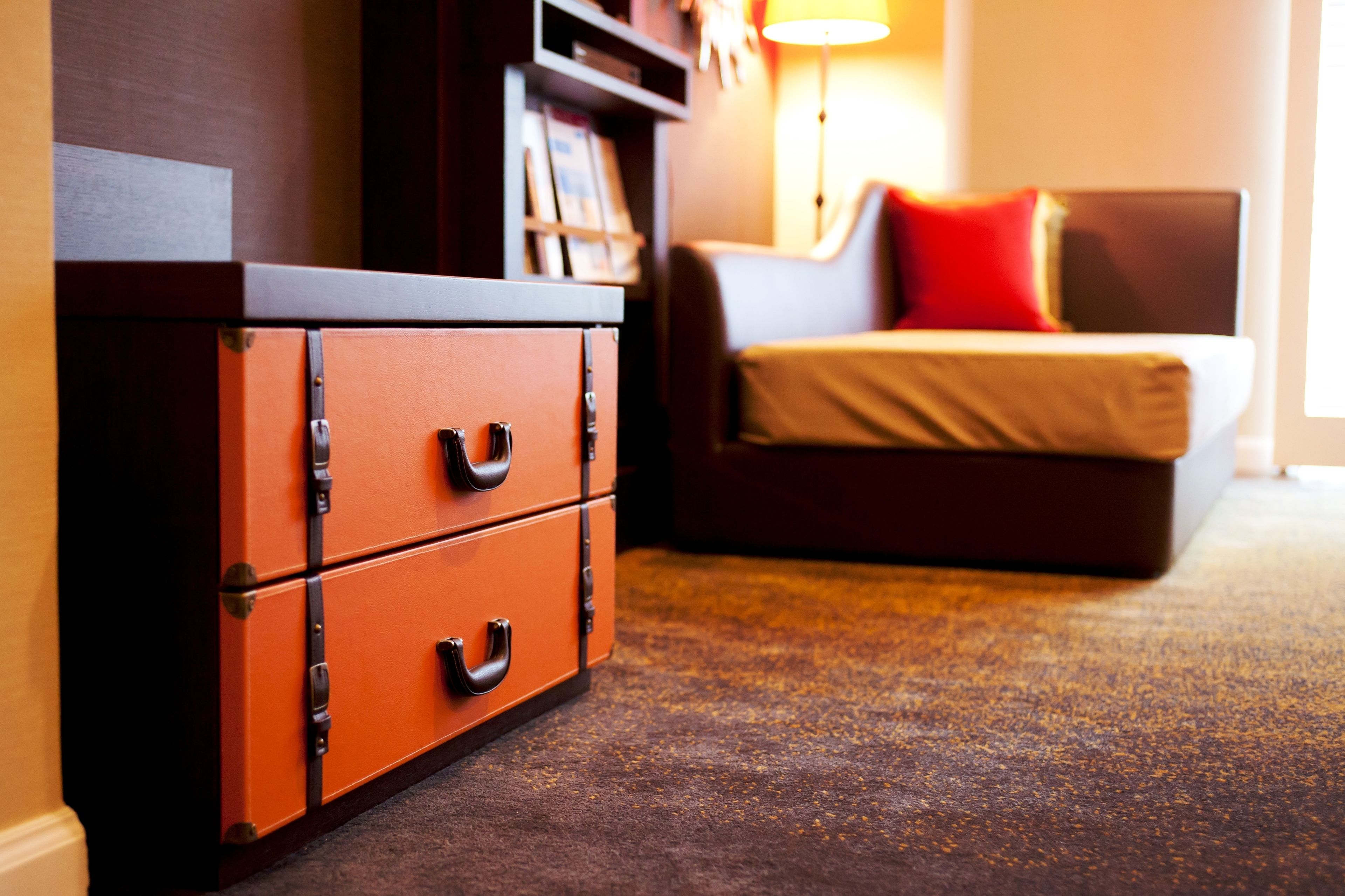 A hotel room with a wooden dresser, a bed with a red pillow, and a lamp.